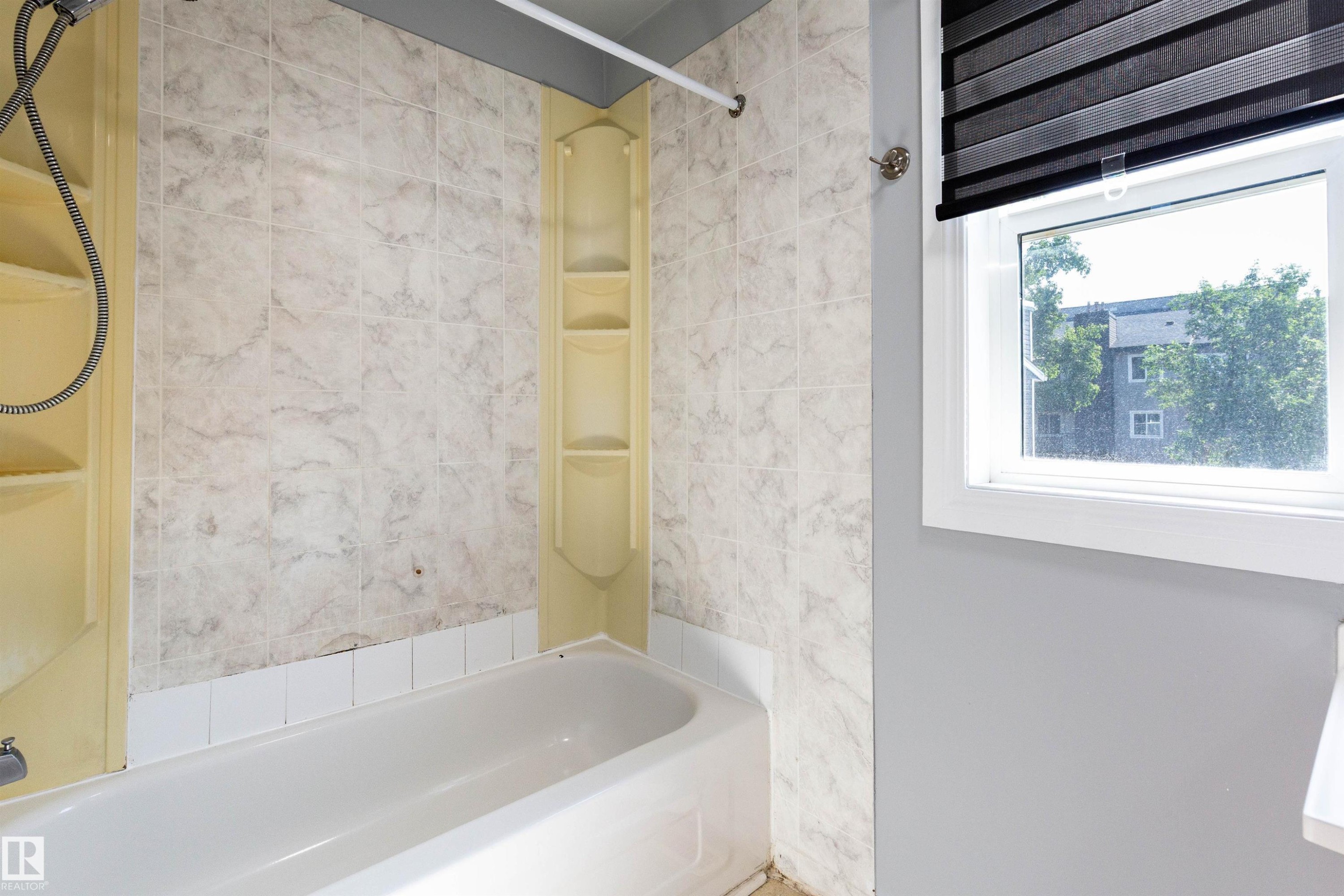 White porcelain bathtub with integrated shelving, surrounded by beige patterned wall tiles and a white window frame with dark roller blinds - 547 Dunluce Road, Edmonton, AB - Indoor Photo Showing Bathroom