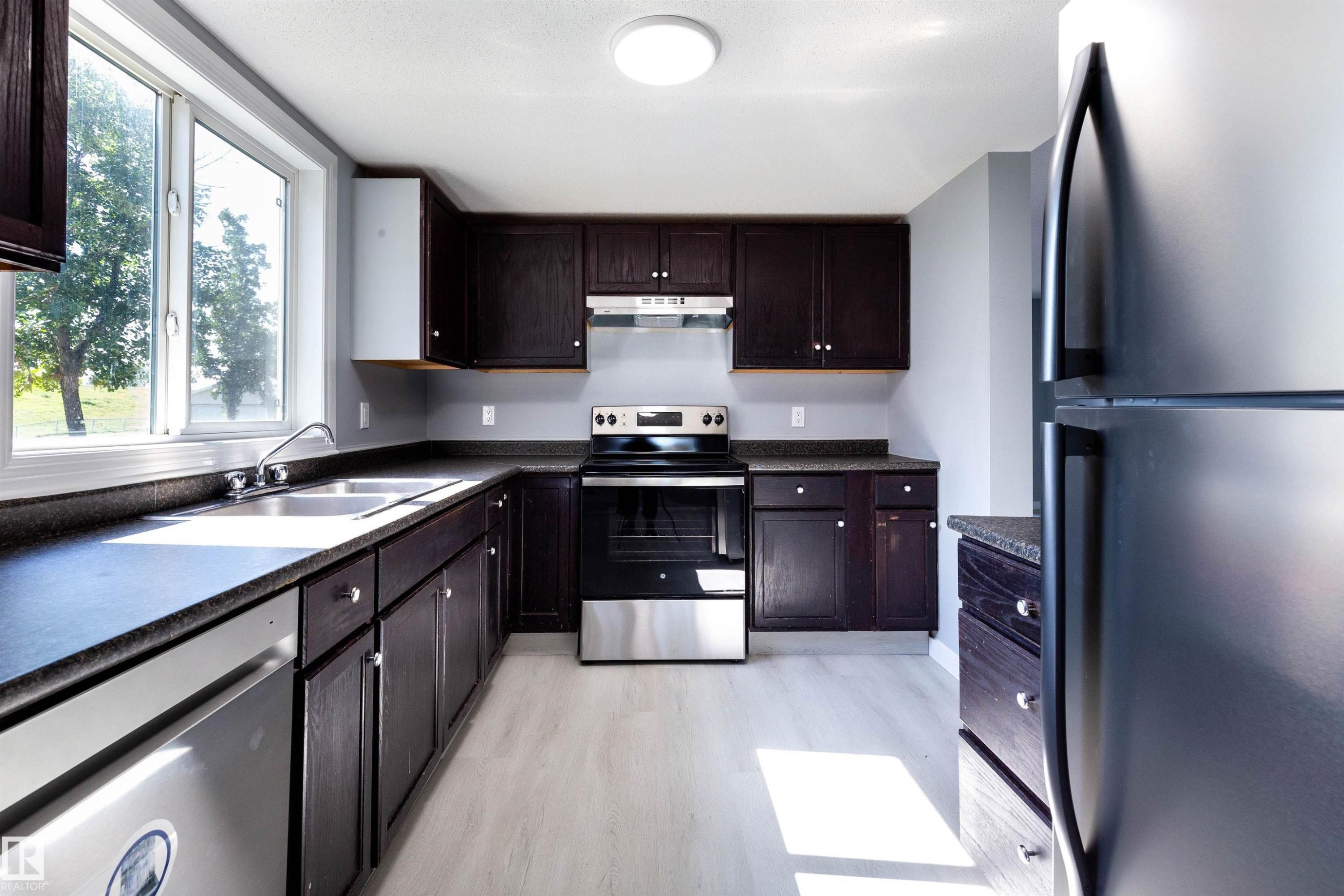 Kitchen featuring dark wood-finish cabinetry, light wood-finish flooring, stainless steel appliances, a double basin sink, and a large window - 547 Dunluce Road, Edmonton, AB - Indoor Photo Showing Kitchen With Stainless Steel Kitchen With Double Sink