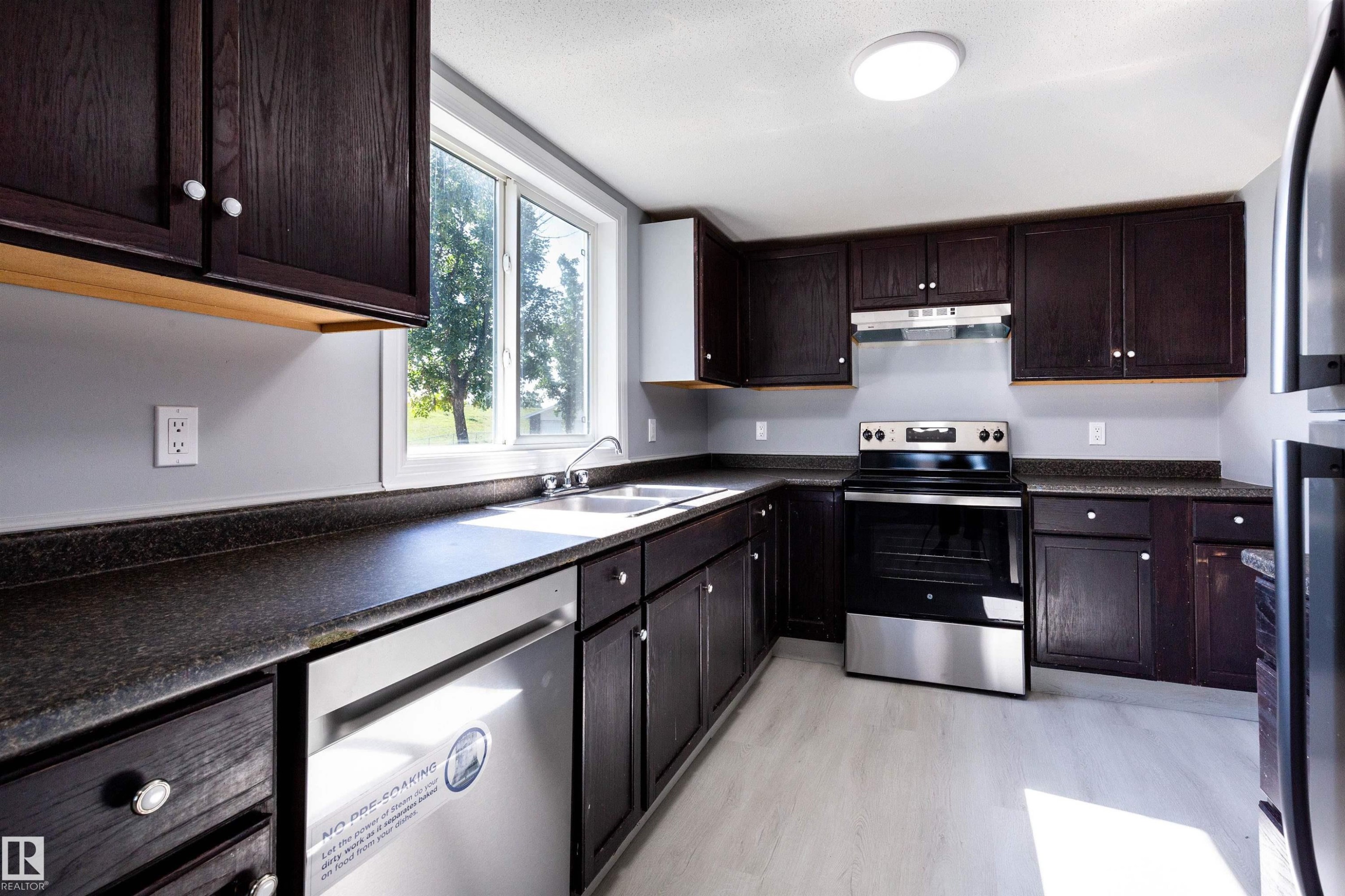 Kitchen featuring dark wood cabinetry, solid surface countertops, stainless steel appliances, a double basin sink, and light wood-finish flooring - 547 Dunluce Road, Edmonton, AB - Indoor Photo Showing Kitchen With Double Sink