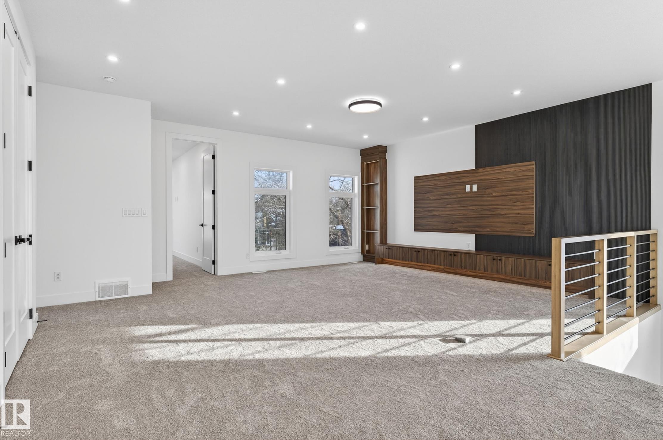 Spacious carpeted room featuring recessed lighting, multiple windows, a built-in media center with wood-finish cabinetry and shelving, and a contemporary railing system with metal balusters - 5820 110 Street, Edmonton, AB - Indoor Photo Showing Other Room
