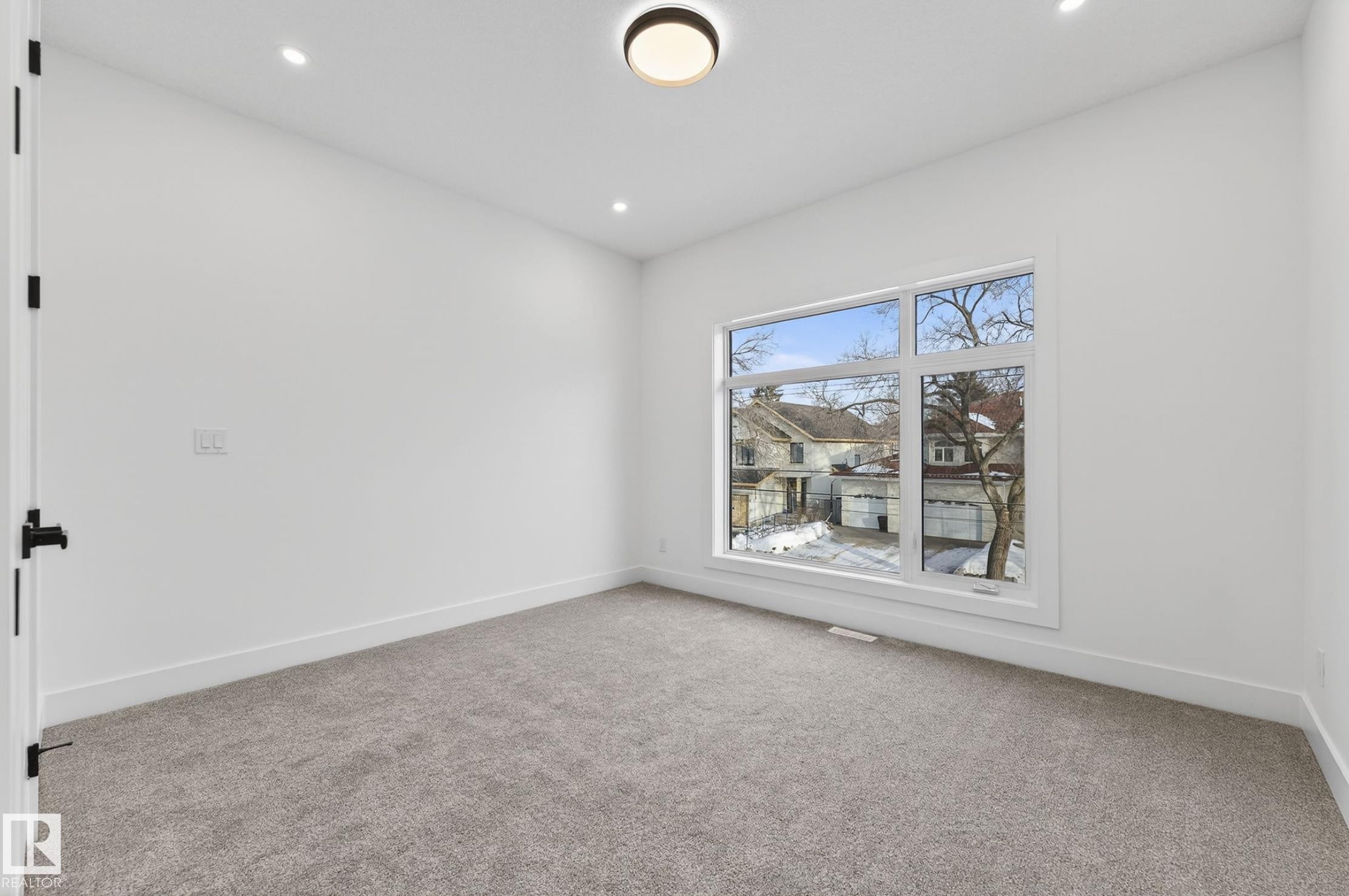 Spacious room featuring a large multi-pane window, recessed lighting, a modern flush-mount ceiling fixture, plush gray carpeting, and crisp white trim - 5820 110 Street, Edmonton, AB - Indoor Photo Showing Other Room