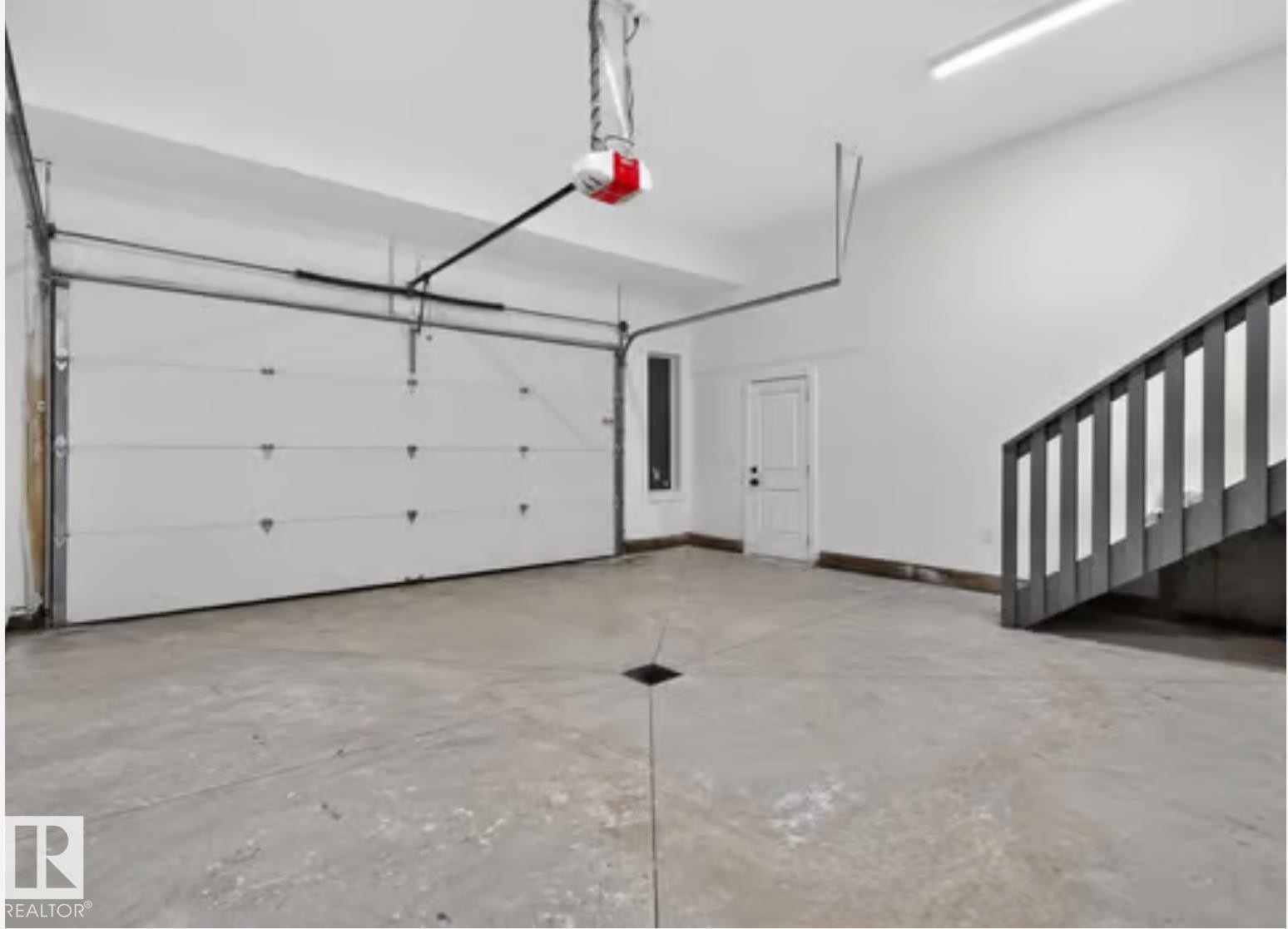 Spacious garage featuring a concrete floor with central drainage, a white paneled garage door with an automatic opener, a white interior door, and a modern staircase with dark wood-finish risers and balusters - 5820 110 Street, Edmonton, AB - Indoor Photo Showing Garage