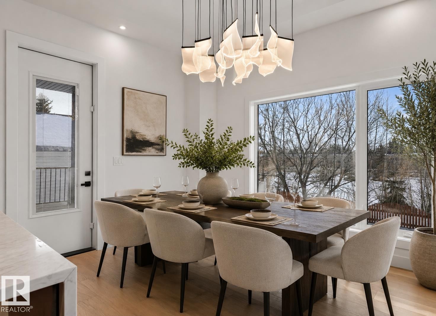 Dining area featuring a contemporary chandelier, expansive windows, a wood-finish dining table, light wood-finish flooring, and a white exterior door with a glass panel - 5820 110 Street, Edmonton, AB - Indoor Photo Showing Dining Room