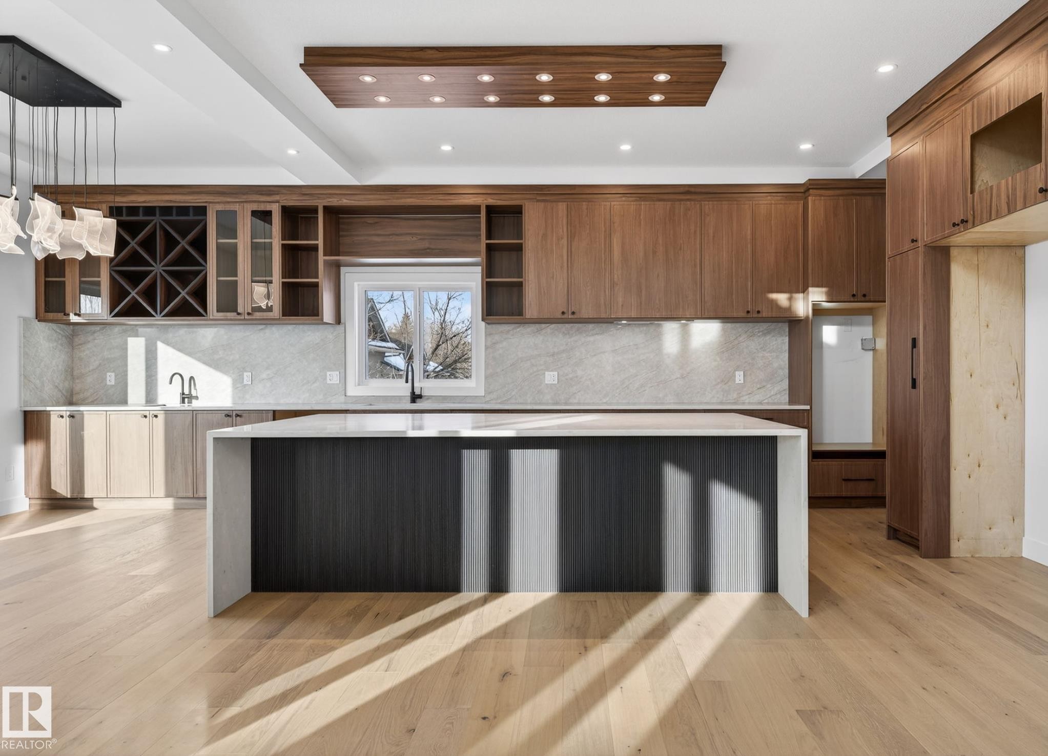 Spacious kitchen featuring a large waterfall island, extensive wood cabinetry, light wood-finish flooring, a contemporary chandelier, and a recessed wood ceiling panel with integrated lighting - 5820 110 Street, Edmonton, AB - Indoor Photo Showing Kitchen With Upgraded Kitchen