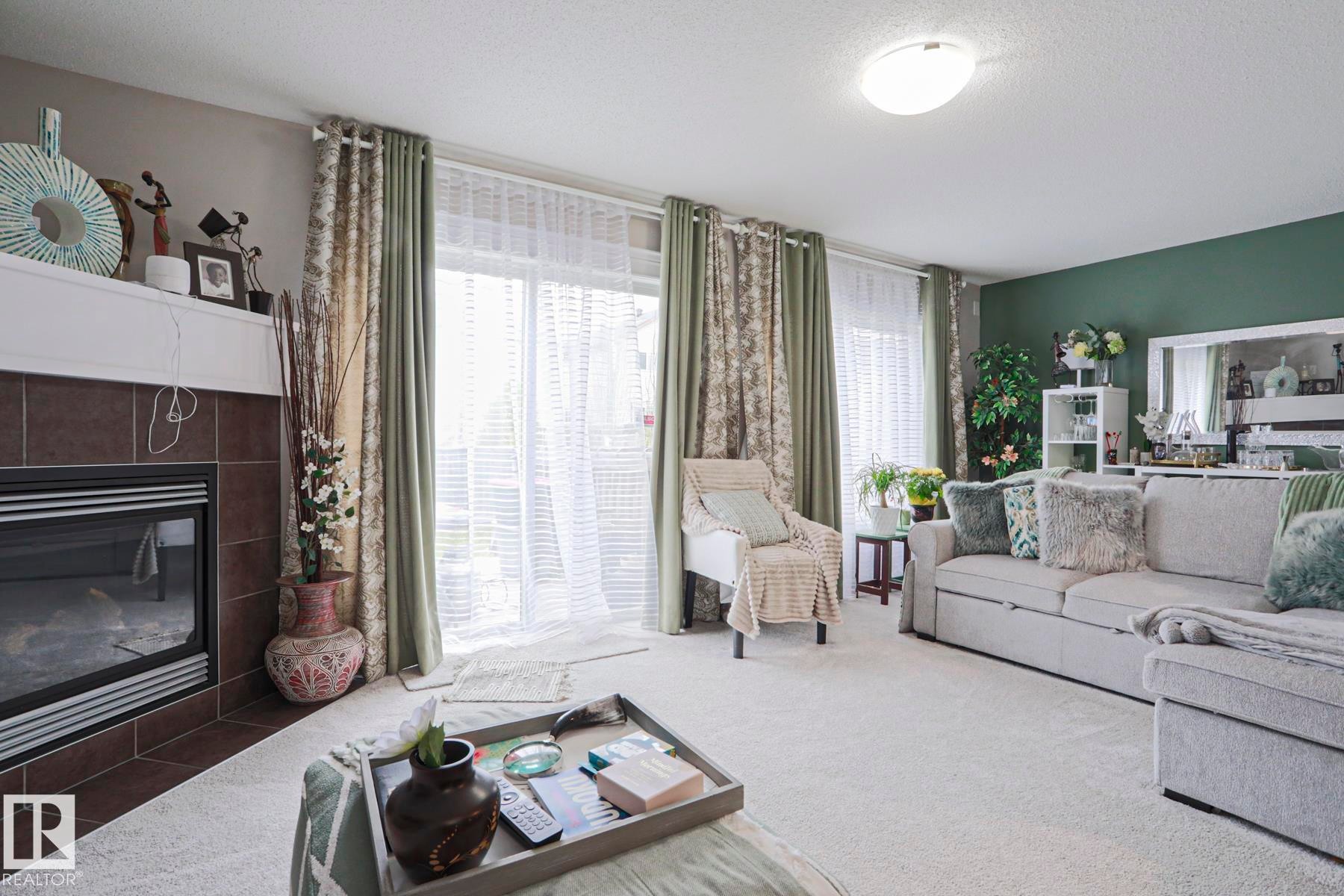 Living area featuring a fireplace with a tiled surround and white mantel, a sliding glass door with sheer and patterned drapes, a large light-toned sectional sofa, and light-colored carpeting throughout - 1621 64 Street, Edmonton, AB - Indoor Photo Showing Living Room With Fireplace