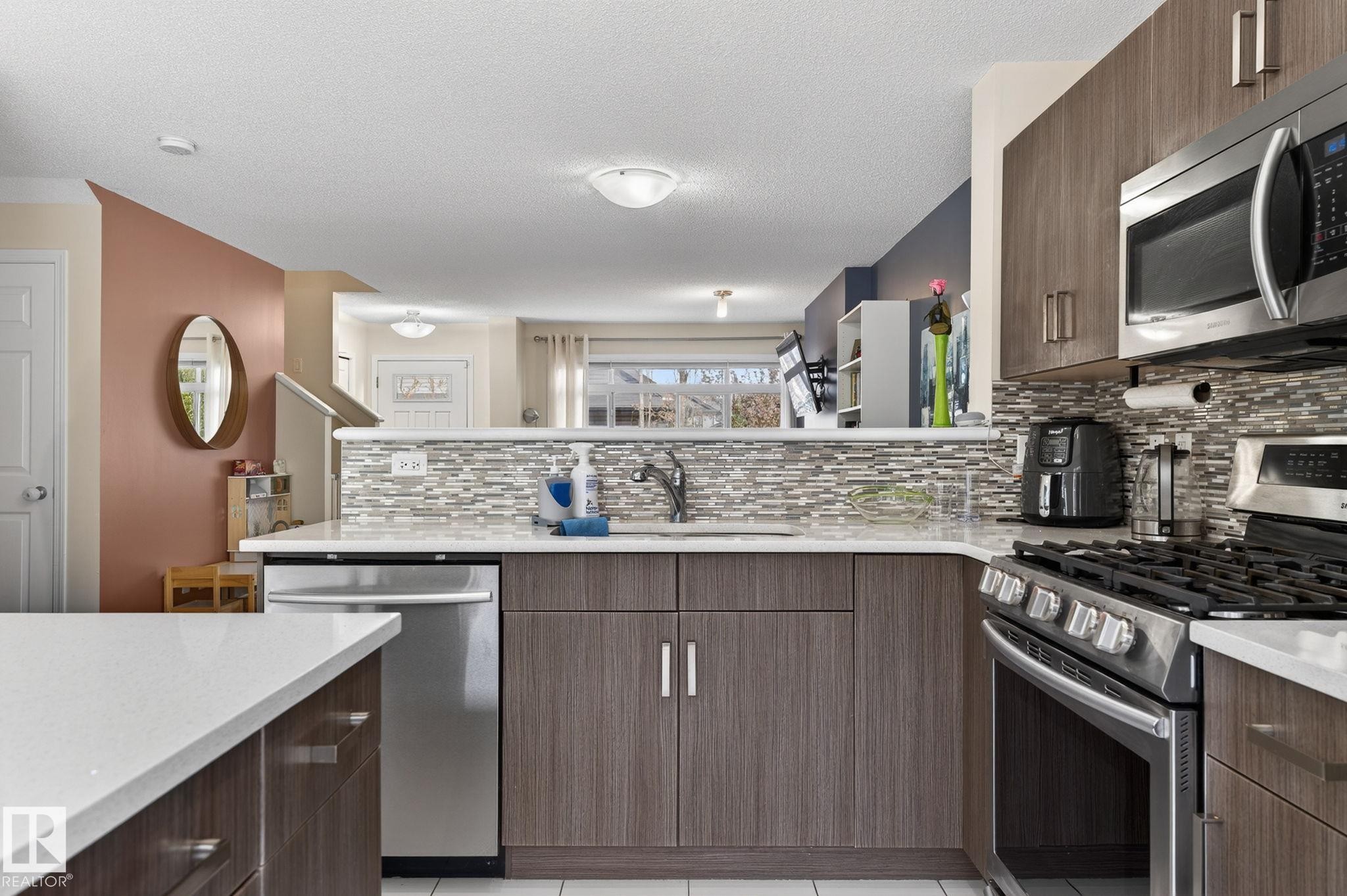 Modern kitchen featuring wood-finish cabinetry, stainless steel appliances, and a mosaic tile backsplash - 16 7503 Getty Gate, Edmonton, AB - Indoor Photo Showing Kitchen With Upgraded Kitchen