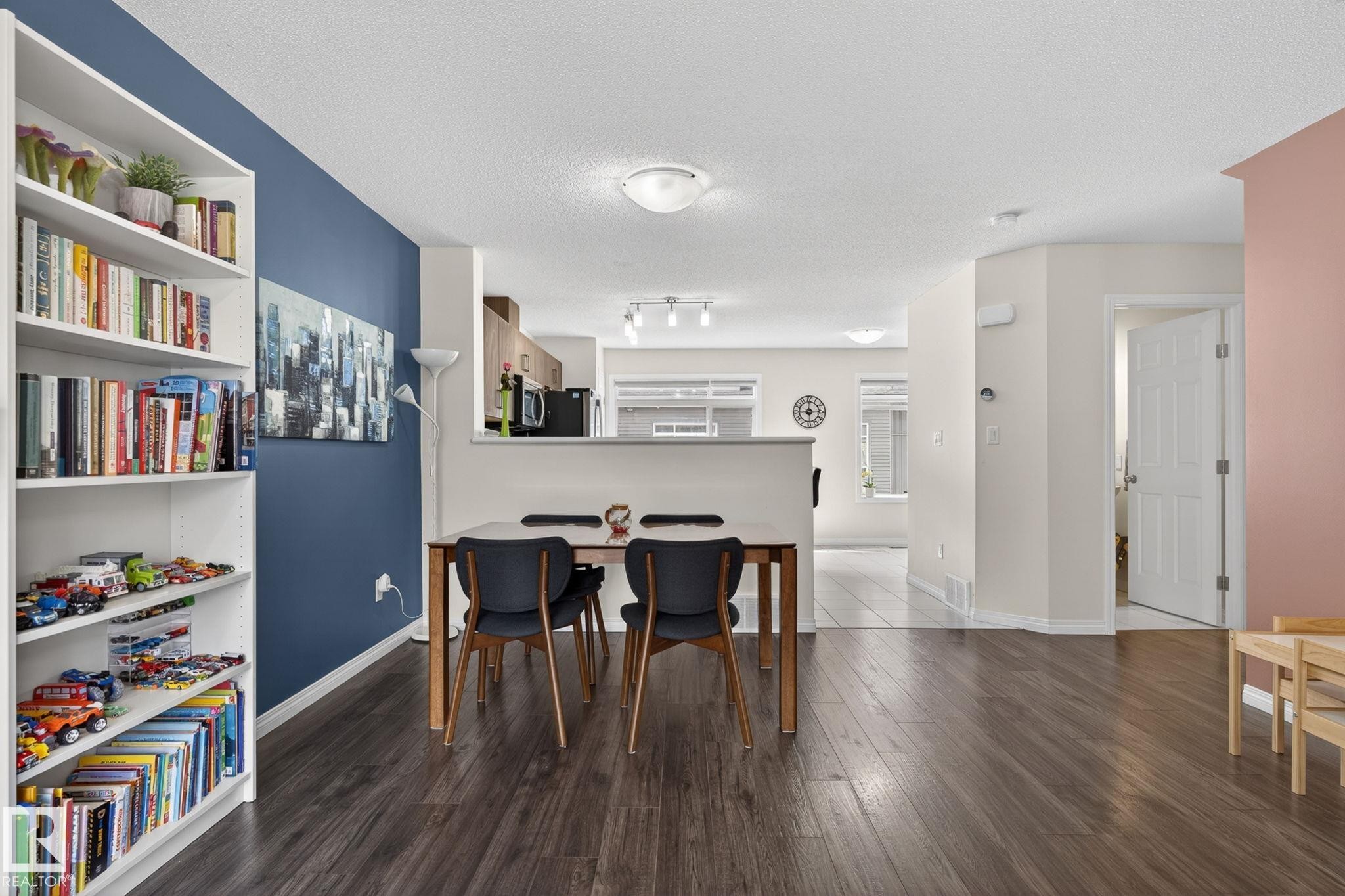 Spacious open-concept living area featuring wood-finish flooring, a built-in white bookshelf, and a half-wall partition - 16 7503 Getty Gate, Edmonton, AB - Indoor