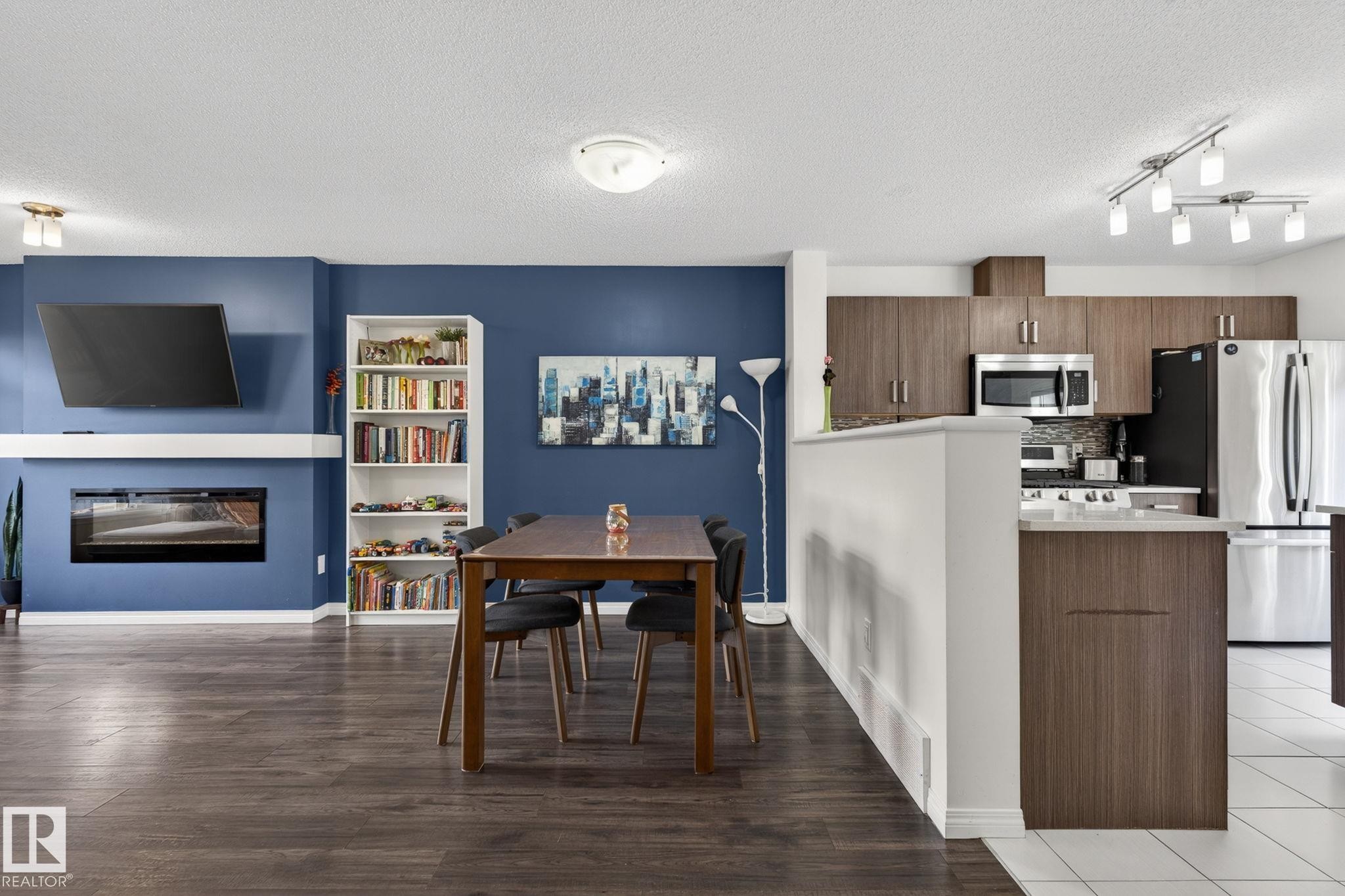 Spacious open-concept living area featuring wood-finish flooring, a built-in electric fireplace with white mantel, a white built-in bookshelf, and a kitchen with stainless steel appliances, dark wood-finish cabinetry, and track lighting - 16 7503 Getty Gate, Edmonton, AB - Indoor Photo Showing Kitchen