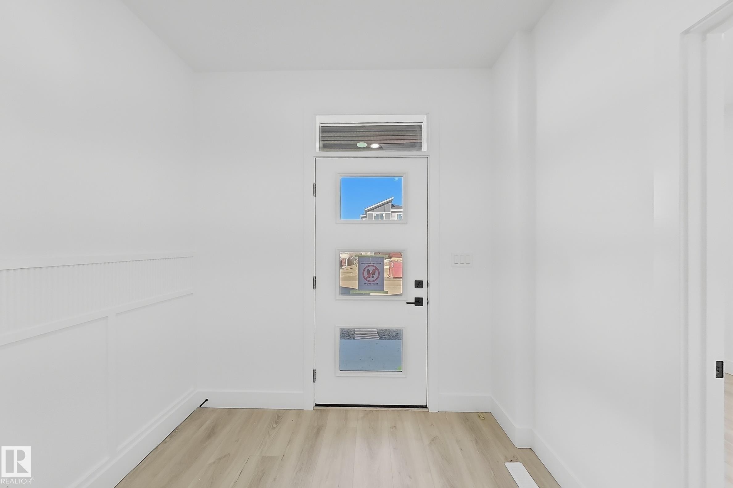 Entryway featuring a white door with three glass inserts, an overhead transom window, light wood-finish flooring, white baseboards, and wainscoting on the left wall - 4215 63 St, Beaumont, AB - Indoor Photo Showing Other Room