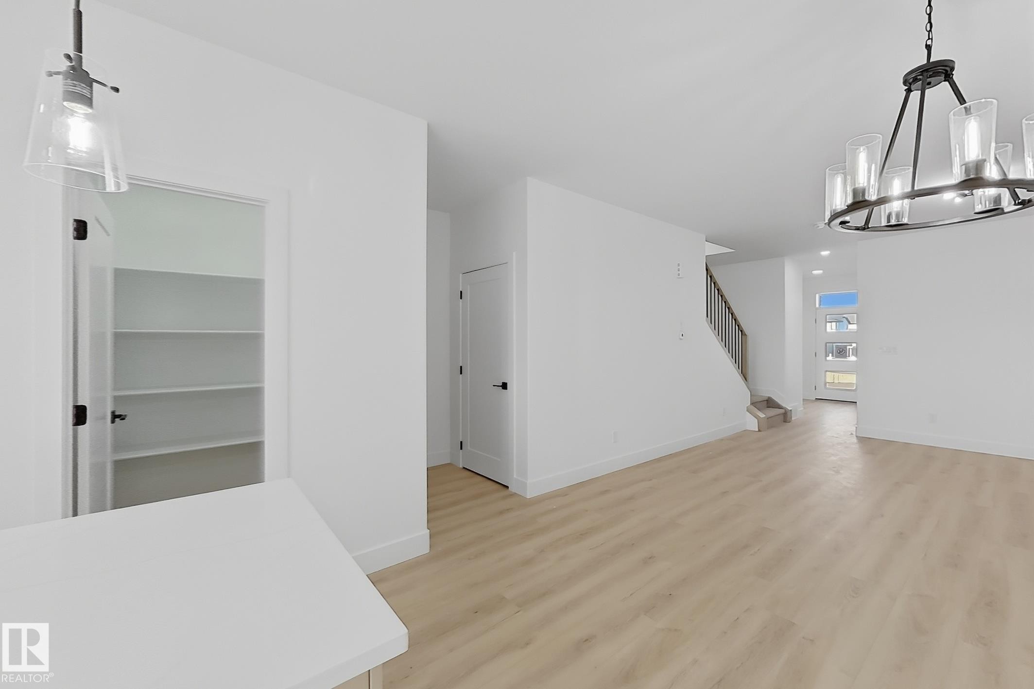 Open-concept living area featuring light wood-finish flooring, a contemporary chandelier, a staircase with dark railings, a white paneled entry door with frosted glass inserts, and a pantry with built-in shelving and a dark hardware door - 4215 63 St, Beaumont, AB - Indoor Photo Showing Other Room