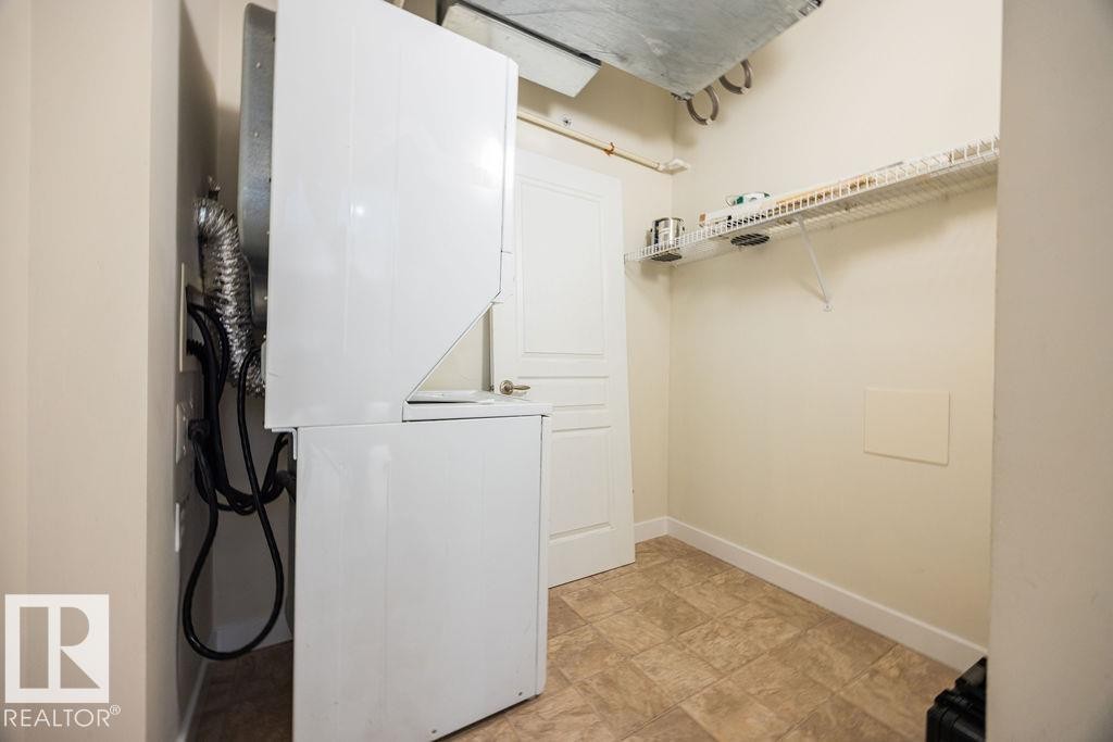 Dedicated laundry area featuring a white stackable washer and dryer unit, complemented by a wire shelving system and a tiled floor - #214 10333 112 St Nw, Edmonton, AB - Indoor