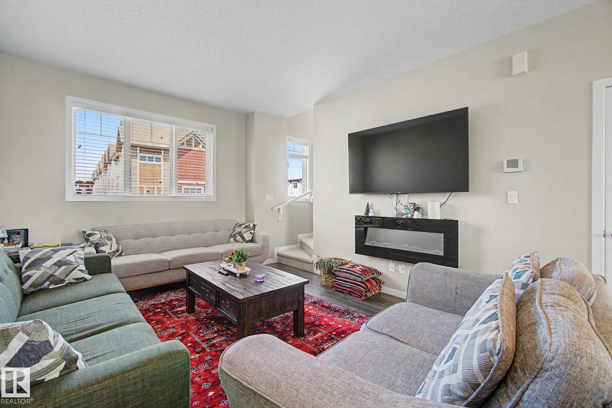 Spacious living area featuring light-colored walls, wood-finish flooring, and a large window with blinds - 17 320 Secord Boulevard, Edmonton, AB - Indoor Photo Showing Living Room With Fireplace