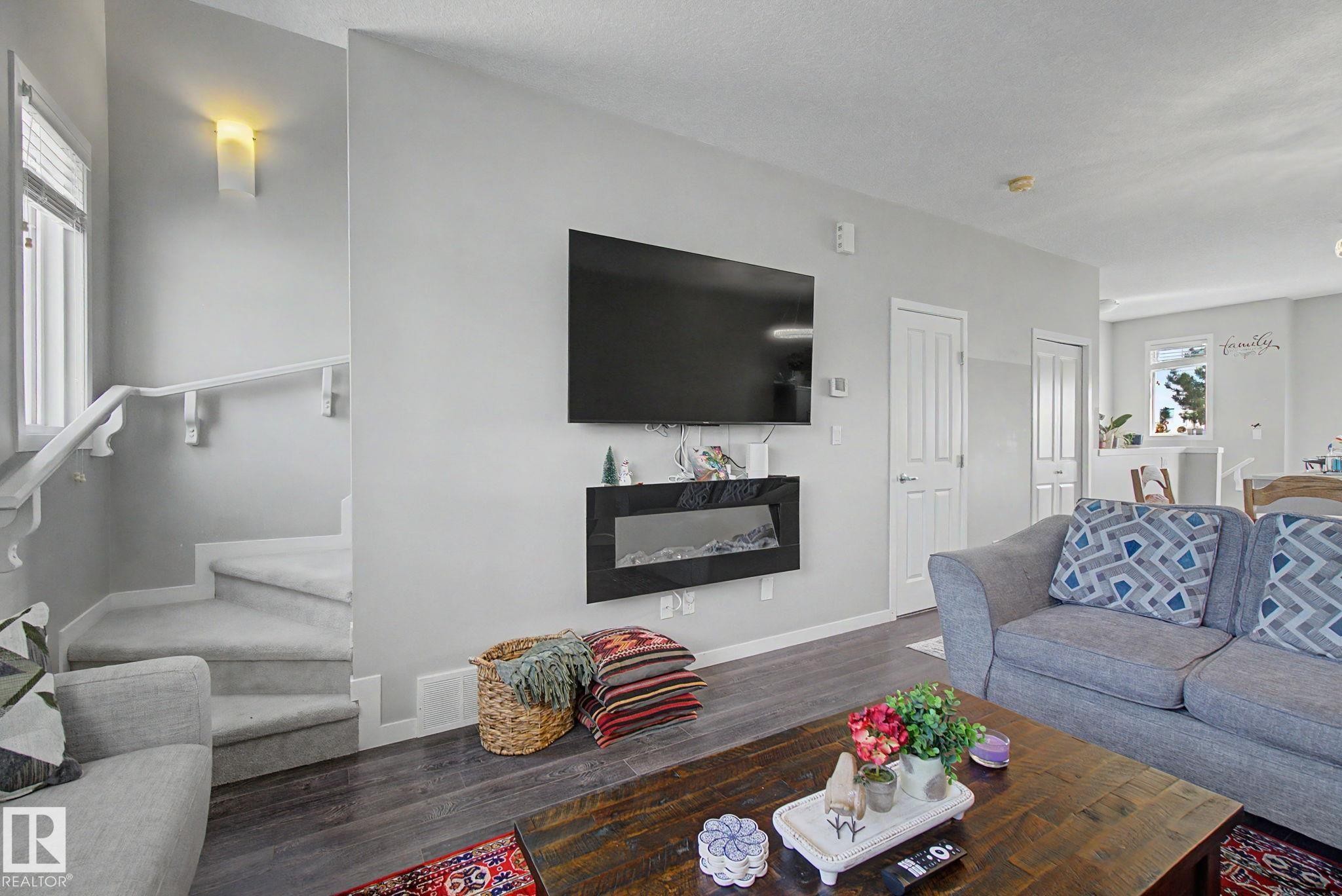 Living area featuring wood-finish flooring, a wall-mounted electric fireplace, and a staircase with carpeted treads and a white handrail - 17 320 Secord Boulevard, Edmonton, AB - Indoor Photo Showing Living Room With Fireplace