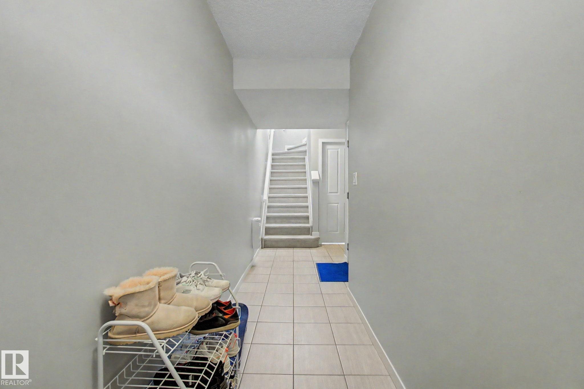 Hallway featuring light-toned tile flooring, light gray painted walls, and a staircase with light-toned carpeted treads - 17 320 Secord Boulevard, Edmonton, AB - Indoor Photo Showing Other Room