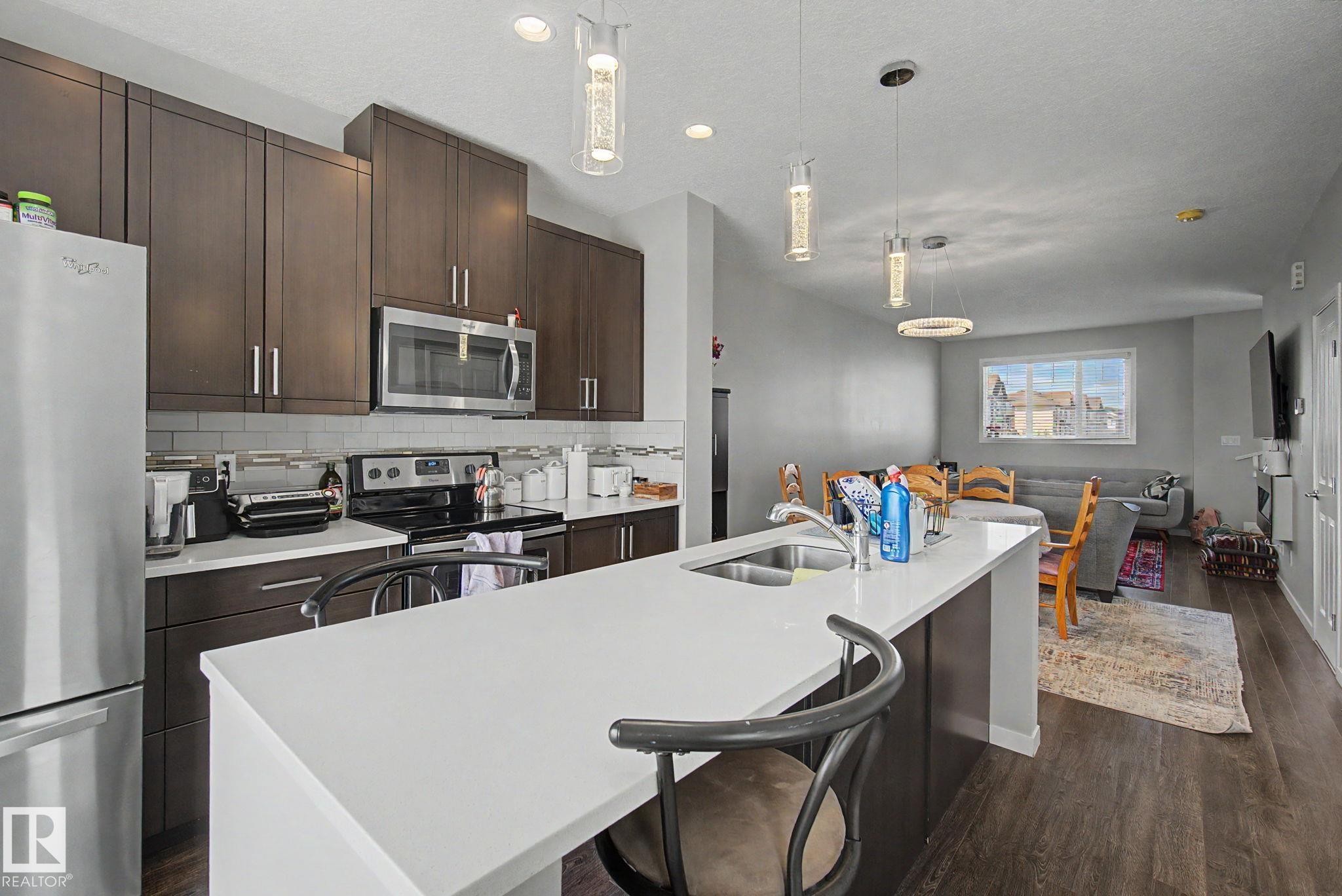 Modern kitchen featuring a white countertop island with an undermount sink, dark wood-finish cabinetry, stainless steel appliances, and a subway tile backsplash - 17 320 Secord Boulevard, Edmonton, AB - Indoor Photo Showing Kitchen With Double Sink With Upgraded Kitchen