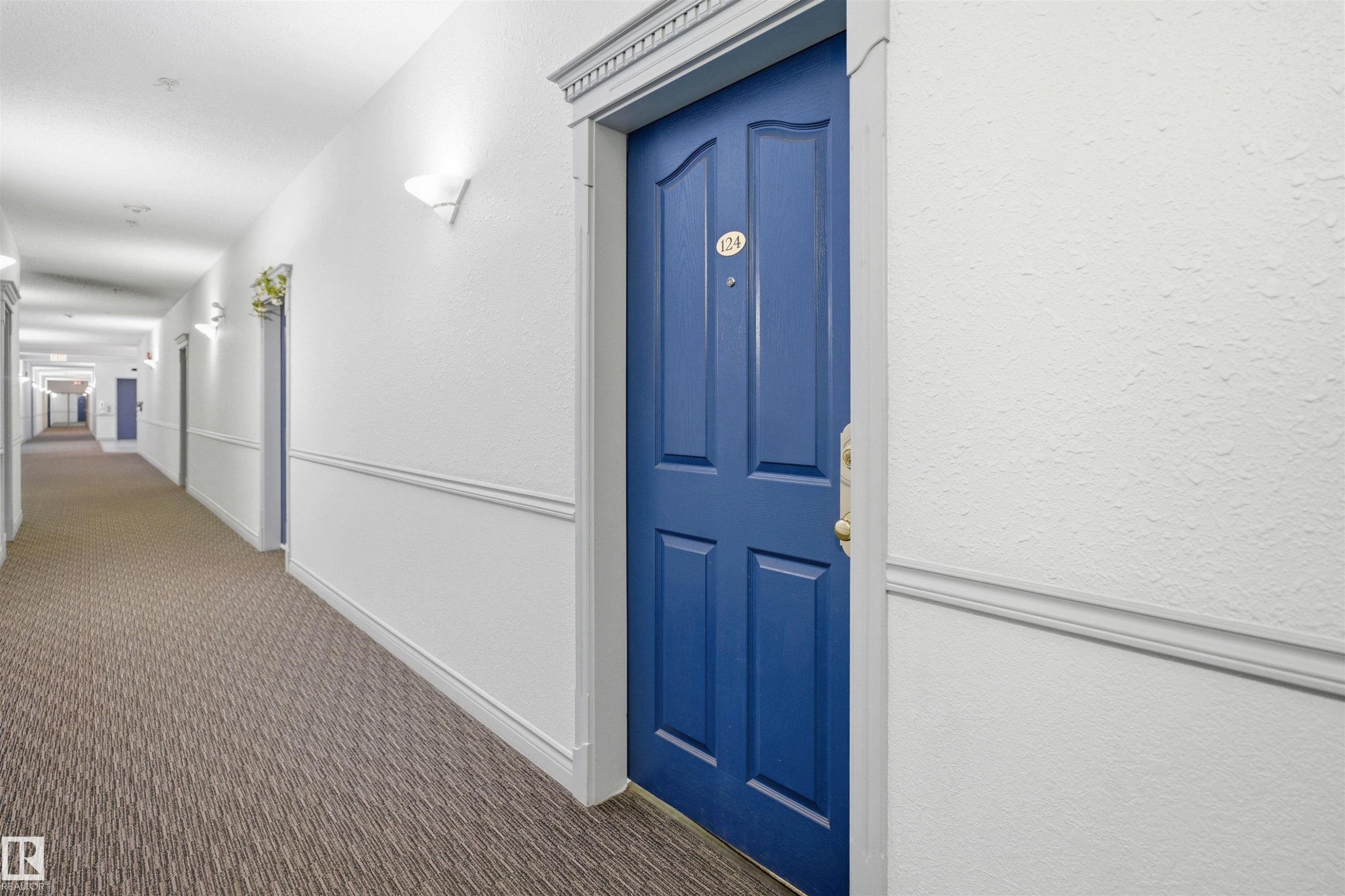 Building corridor featuring a paneled entry door, wall sconces, patterned carpet flooring, and white wainscoting - 124 10511 42 Avenue, Edmonton, AB