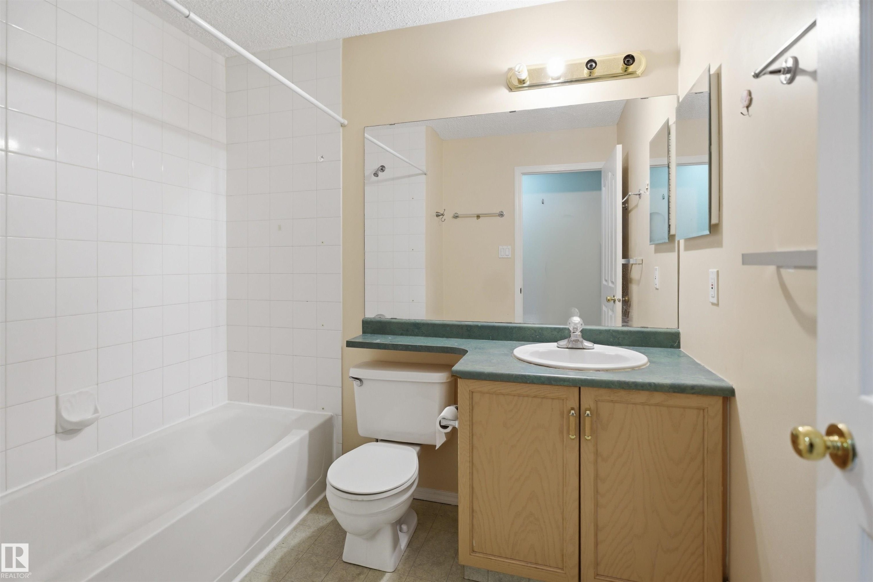 Full bathroom featuring a white tub-shower combination with white tile surround, a vanity with wood-finish cabinetry, a white basin, and a wall-mounted mirror - 124 10511 42 Avenue, Edmonton, AB