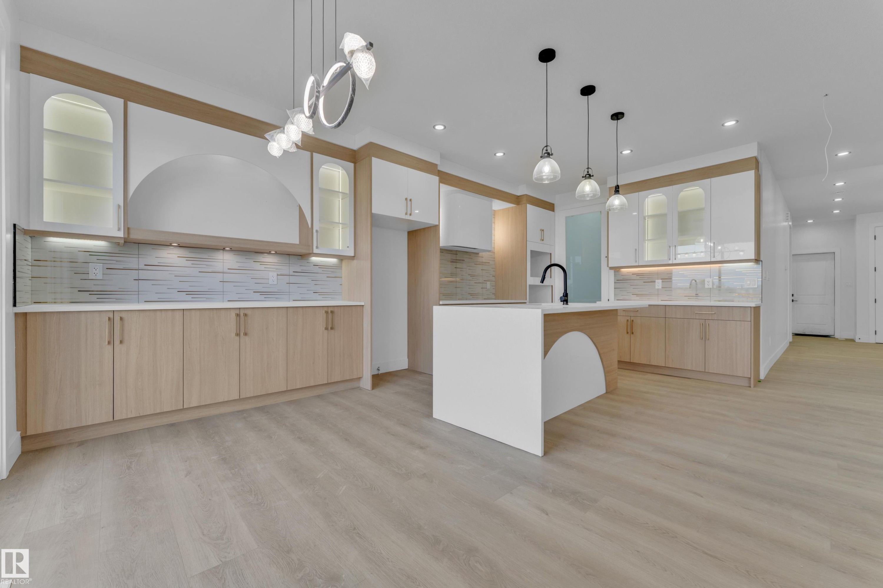 Modern kitchen featuring light wood-finish flooring, two-tone cabinetry with wood accents, a central island with a waterfall countertop, contemporary pendant lighting, and a linear tile backsplash - 449 Crystal Creek Link, Leduc, AB - Indoor Photo Showing Kitchen With Upgraded Kitchen