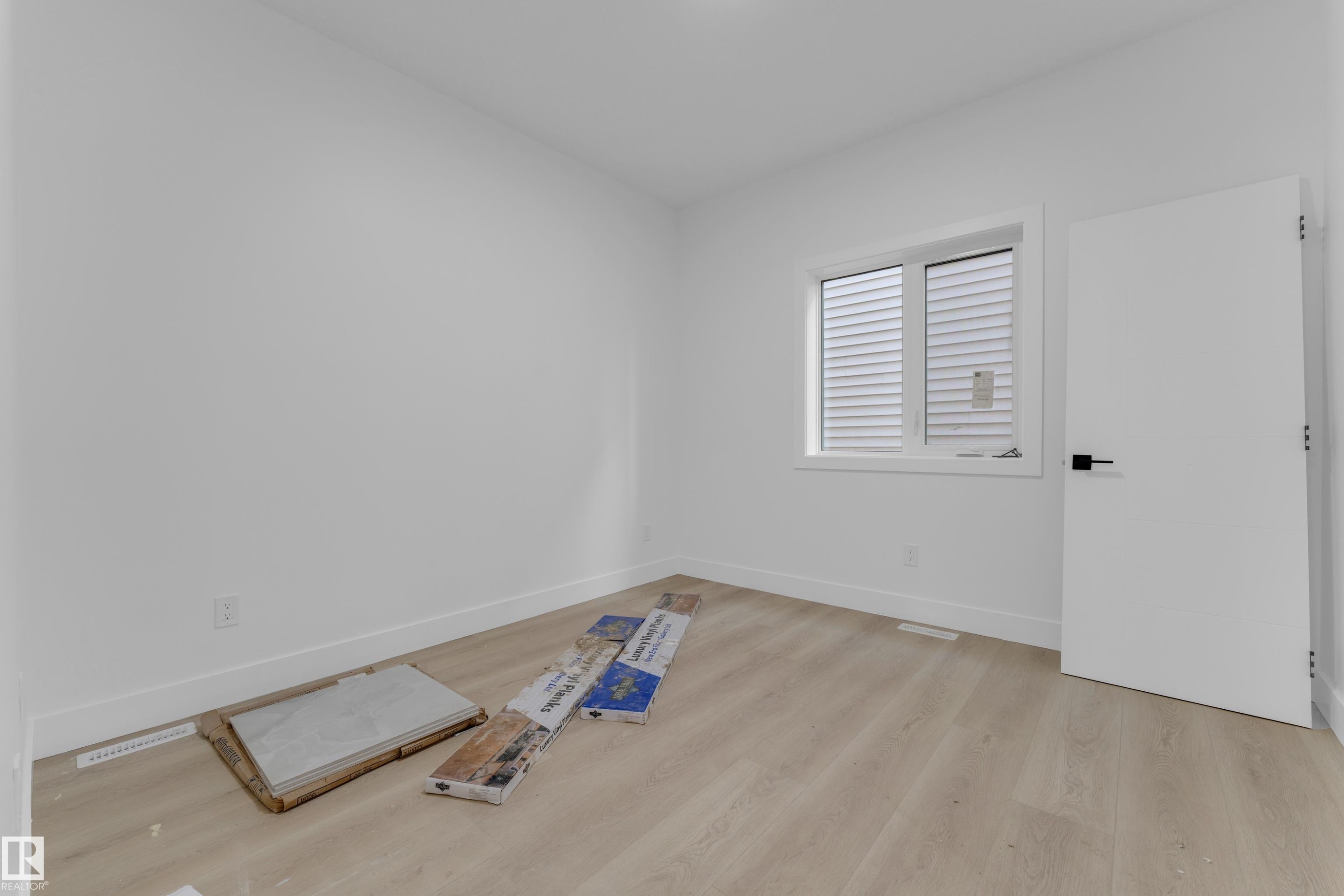 Bright room featuring a large window with white blinds, light wood-finish flooring, white baseboards, and a modern white door with black hardware - 449 Crystal Creek Link, Leduc, AB - Indoor Photo Showing Other Room