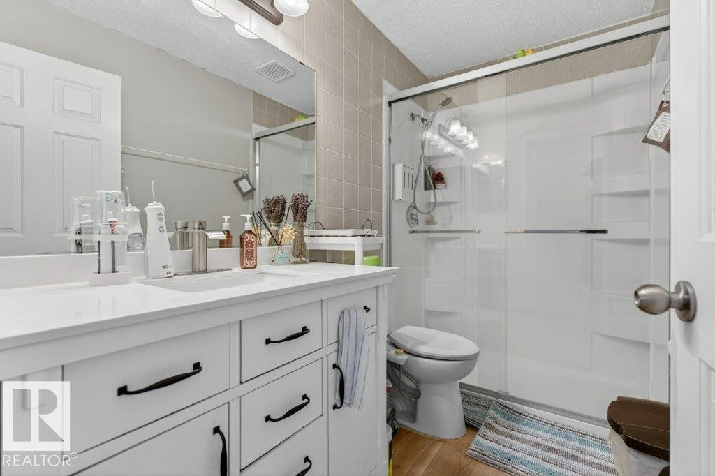 Bathroom featuring a white vanity with black hardware, a solid surface countertop, a large frameless mirror, a shower with a sliding glass door, and wood-finish flooring - 157 Kiniski Crescent, Edmonton, AB - Indoor Photo Showing Bathroom