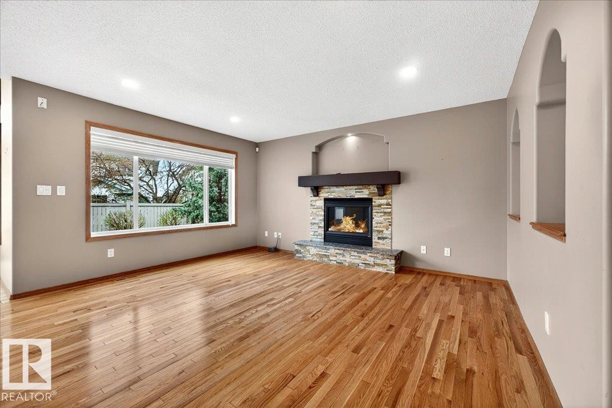 Spacious room featuring wood-finish flooring, a stone-surround fireplace with a wood mantle, and a large window providing outdoor views - 11824 13 Avenue, Edmonton, AB - Indoor Photo Showing Living Room With Fireplace