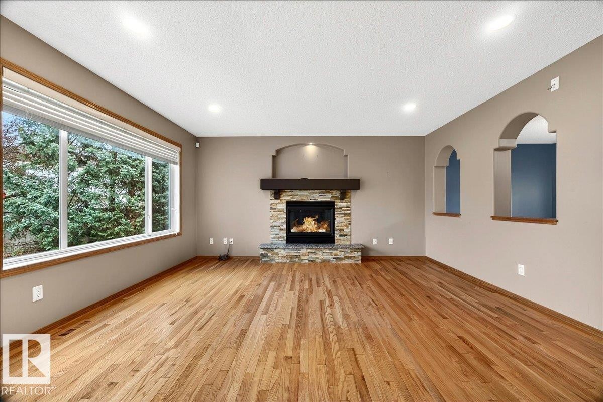 Spacious living area featuring wood-finish flooring, a stone-faced fireplace with a dark wood mantel, recessed lighting, and large windows with natural wood trim - 11824 13 Avenue, Edmonton, AB - Indoor Photo Showing Living Room With Fireplace