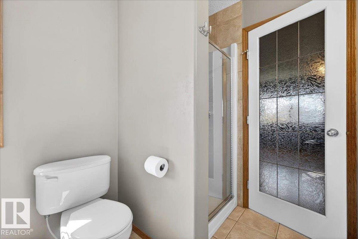 Bathroom featuring white toilet, light-toned wall paint, tiled flooring, and a glass-paneled door with wood trim - 11824 13 Avenue, Edmonton, AB - Indoor Photo Showing Bathroom
