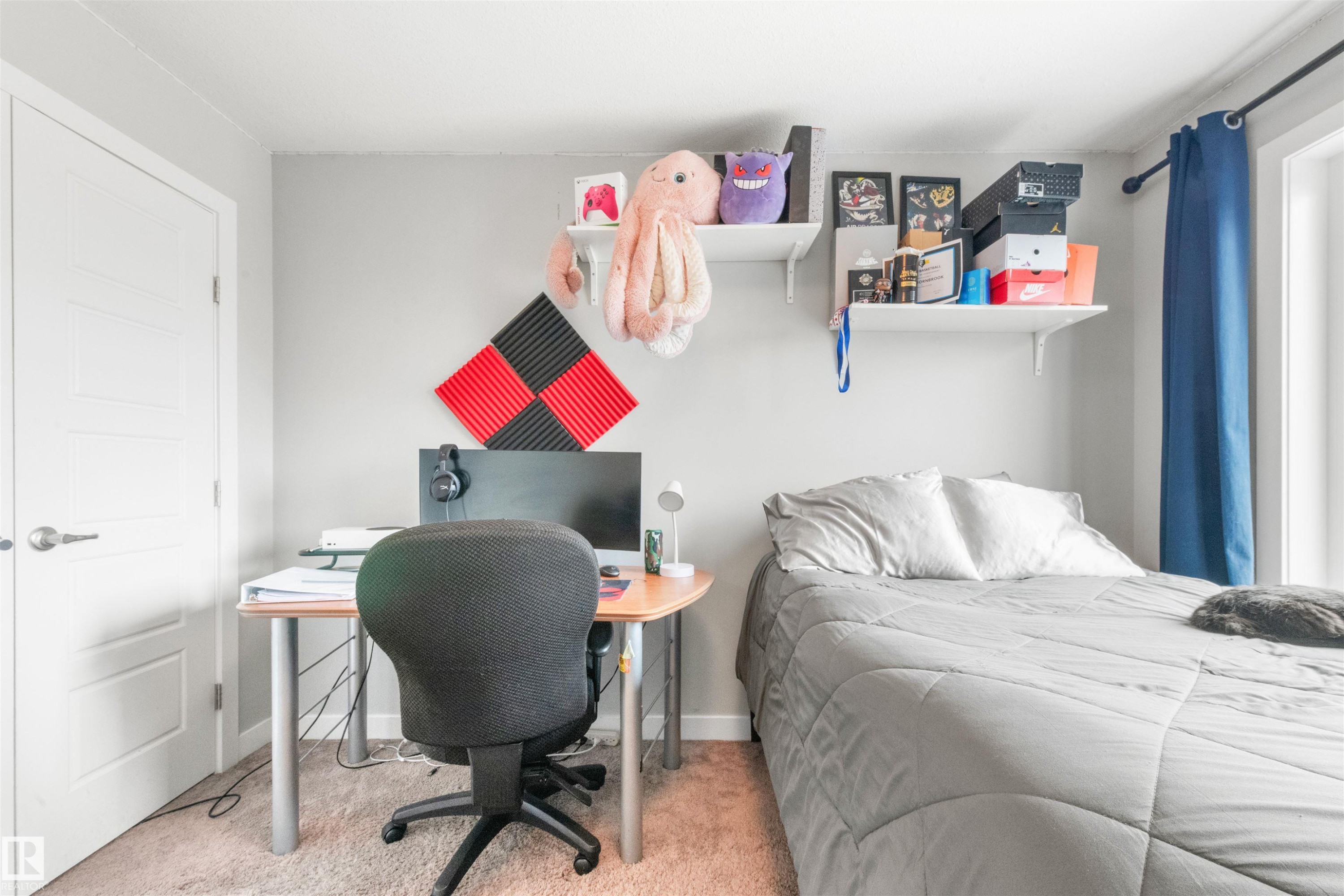 This room features light-colored walls, a bed with a gray comforter, a desk area, and a window with blue curtains - 607 40 Street Sw, Edmonton, AB - Indoor Photo Showing Bedroom