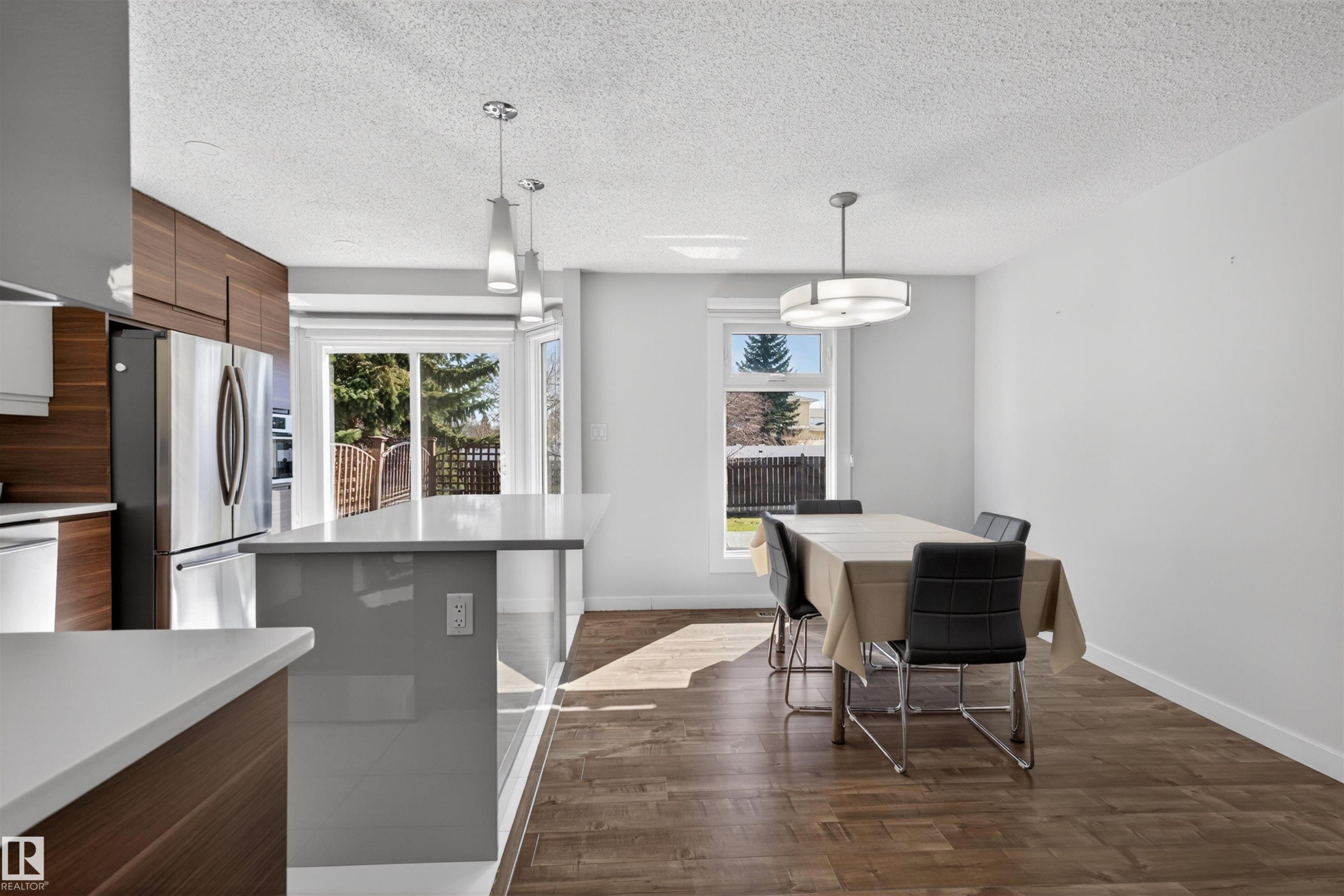 Modern kitchen featuring wood-finish cabinetry, stainless steel refrigerator, white countertops, and a gray island with pendant lighting - 15251 88A Street, Edmonton, AB - Indoor