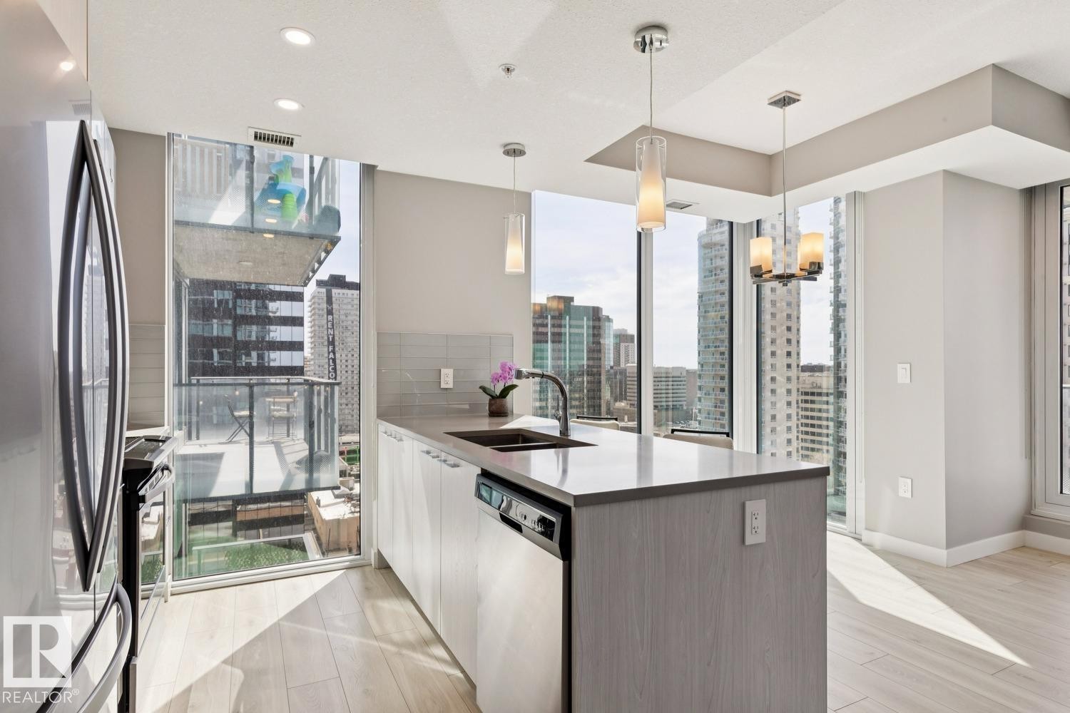 Modern kitchen featuring expansive floor-to-ceiling windows, a central island with an undermount sink, pendant lighting, built-in dishwasher, and light wood-finish flooring - #1601 10180 103 St Nw, Edmonton, AB - Indoor Photo Showing Kitchen With Double Sink With Upgraded Kitchen