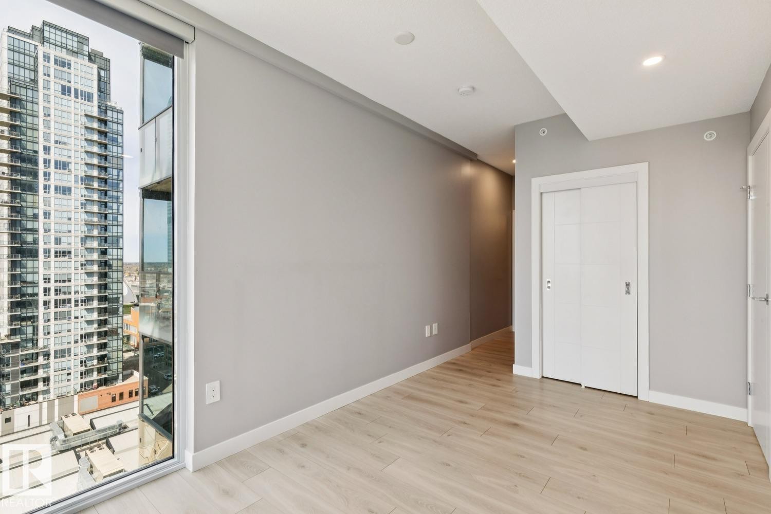 Contemporary interior featuring a wide window, light wood-finish flooring, recessed lighting, a sliding barn door, and a modern city backdrop - #1601 10180 103 St Nw, Edmonton, AB - Indoor Photo Showing Other Room