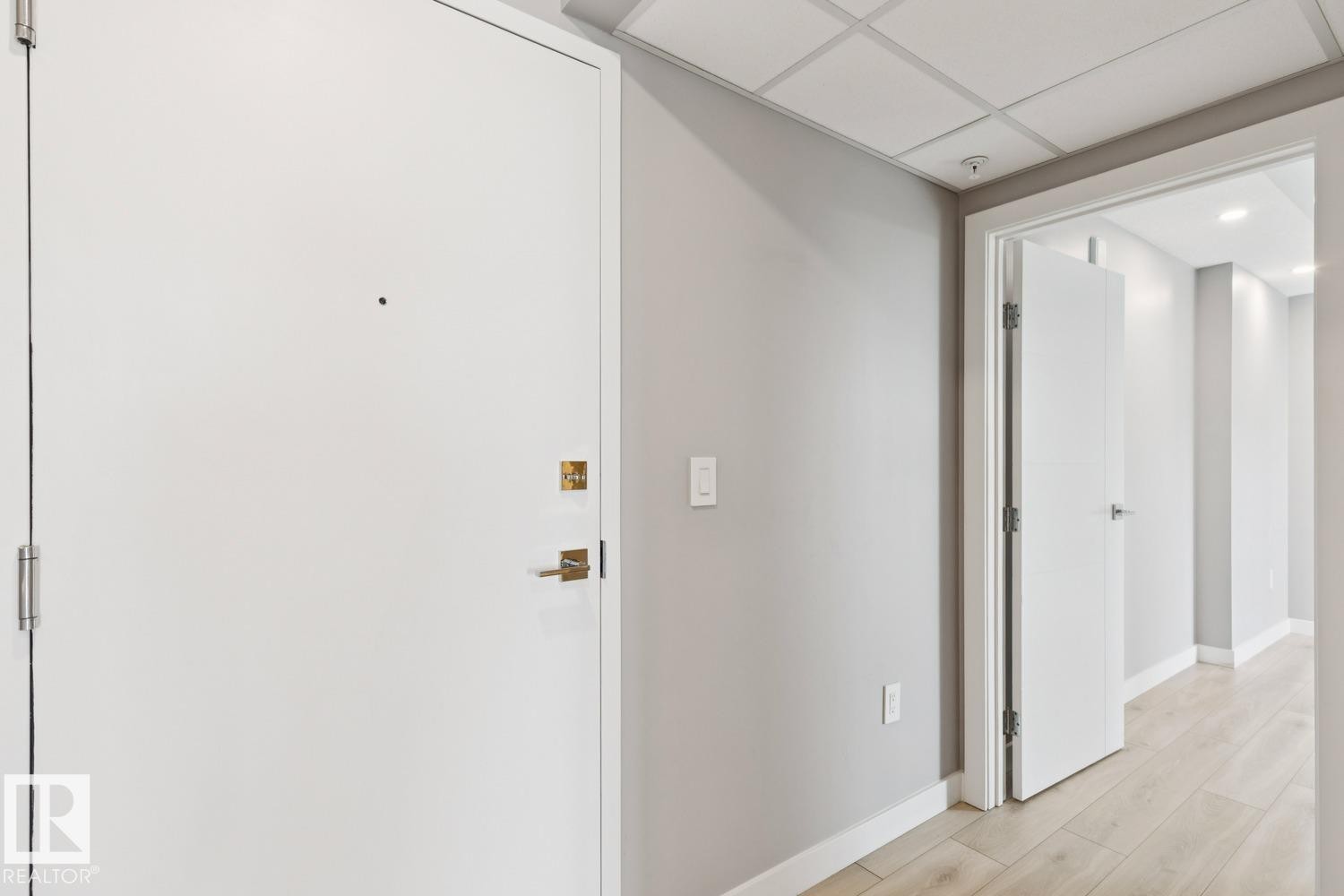 Entryway featuring a white door with brass hardware, light gray painted walls, a dropped ceiling, and light wood-finish flooring - #1601 10180 103 St Nw, Edmonton, AB - Indoor Photo Showing Other Room