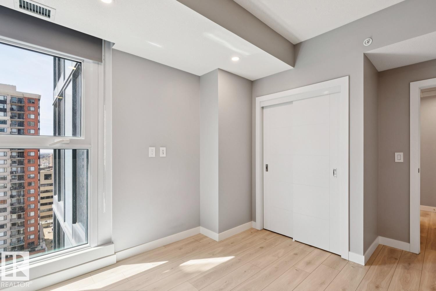 Bright interior space featuring a large window with city buildings visible, light wood-finish flooring, recessed ceiling lighting, a white sliding closet door, and neutral gray wall tones - #1601 10180 103 St Nw, Edmonton, AB - Indoor Photo Showing Other Room