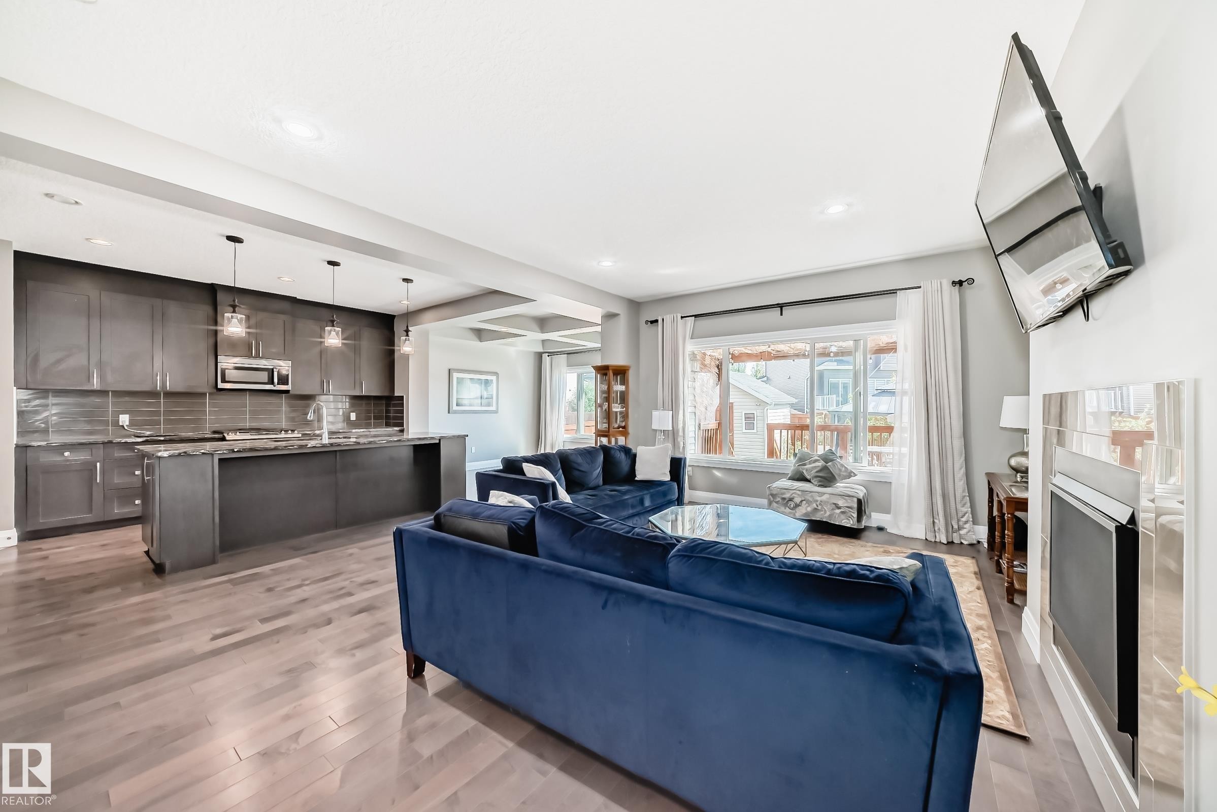 Open-concept living space featuring wood-finish flooring, a contemporary kitchen with dark cabinetry and pendant lighting, a recessed fireplace with a tiled surround, and large windows - 4155 Charles Link, Edmonton, AB - Indoor With Fireplace