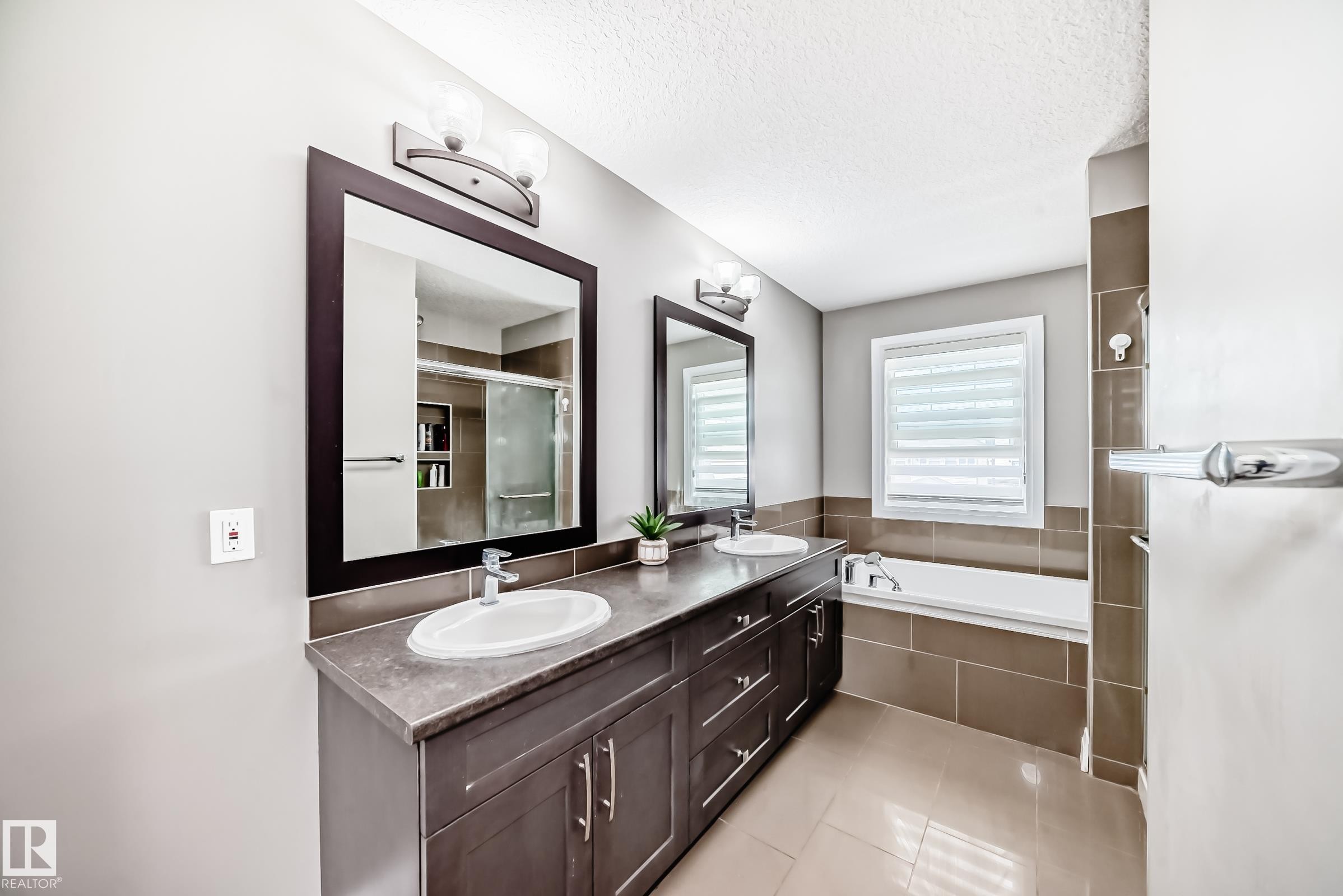 Bathroom featuring a dual vanity with dark wood-finish cabinetry, a built-in tub with tile surround, a large framed mirror, and tile flooring - 4155 Charles Link, Edmonton, AB - Indoor Photo Showing Bathroom