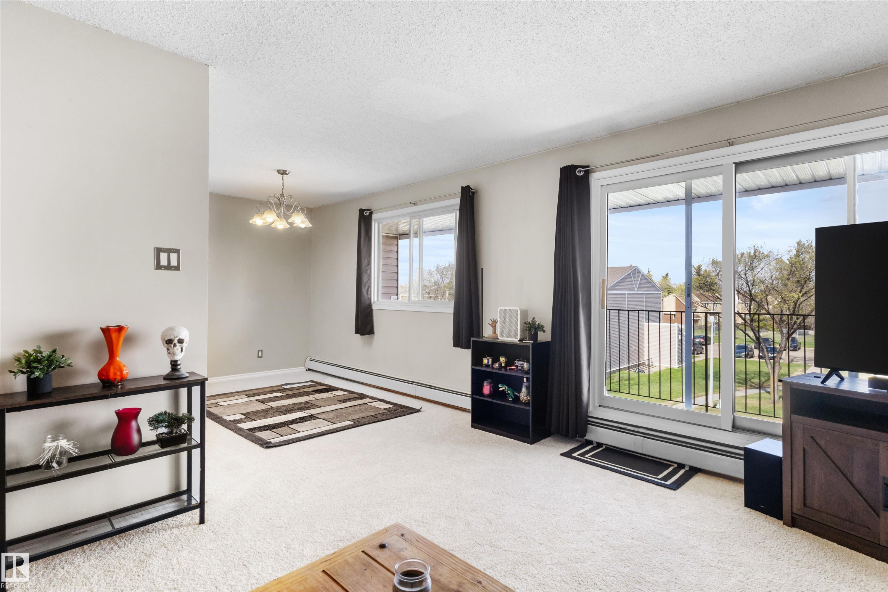 Living area featuring light-colored carpeting, a wall-mounted chandelier, and a sliding glass door providing access to an exterior balcony - 6 3803 76 Street, Edmonton, AB - Indoor