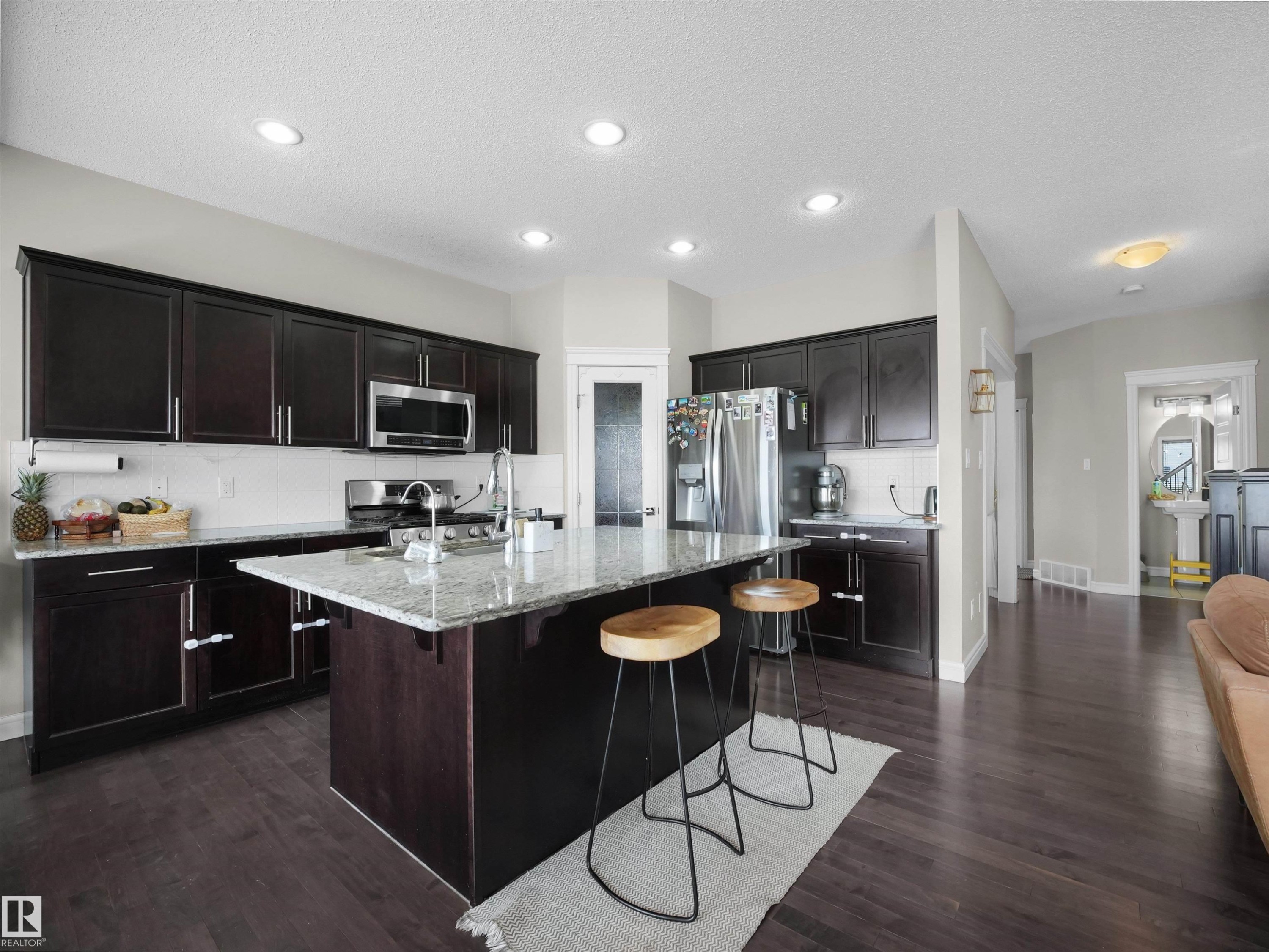 Kitchen featuring dark wood cabinetry, stainless steel appliances, a white tile backsplash, and a large central island with a granite countertop and sink - 3247 15 Avenue Nw, Edmonton, AB - Indoor Photo Showing Kitchen With Upgraded Kitchen