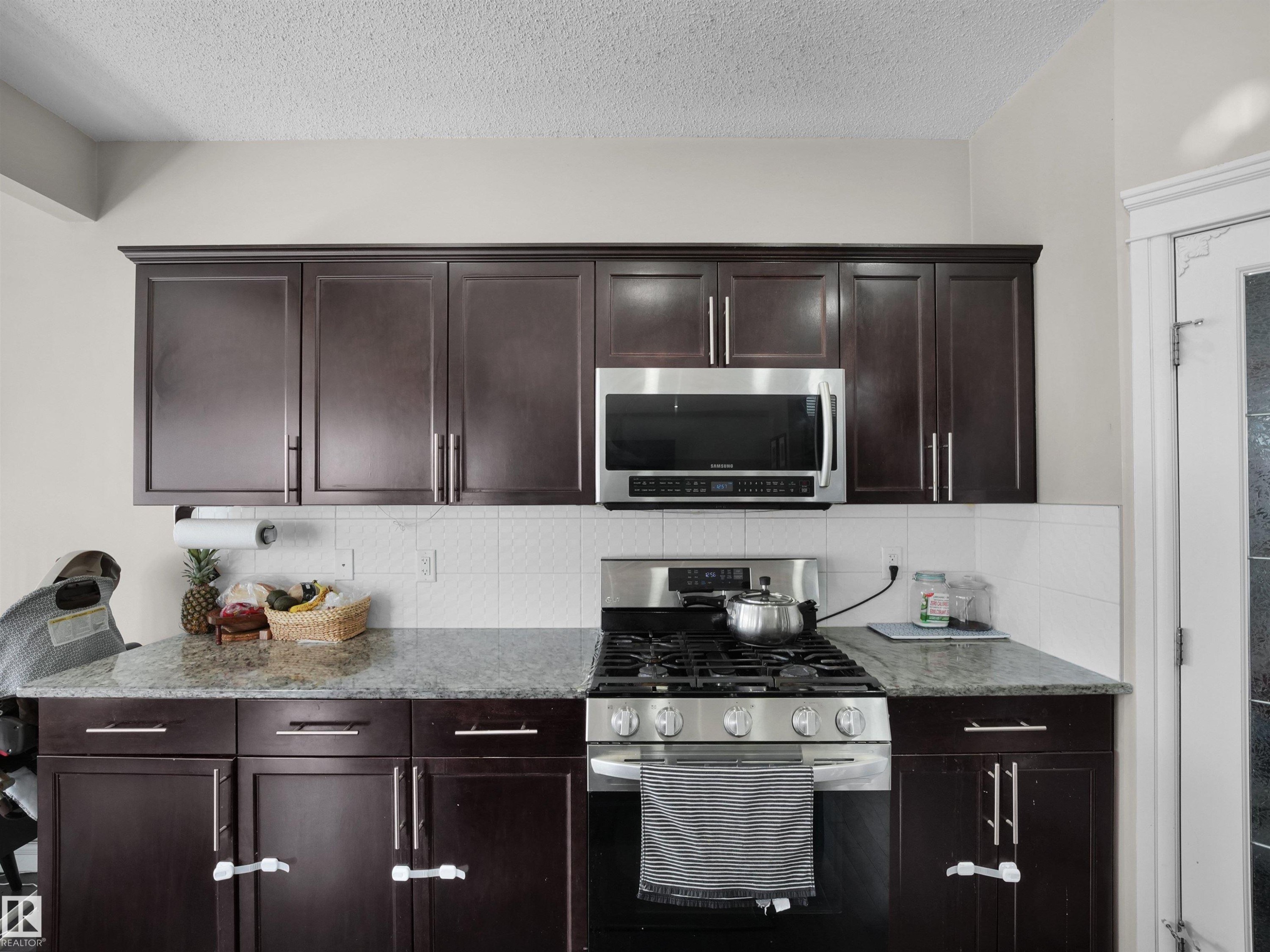 Kitchen featuring dark wood cabinetry, light-toned granite countertops, a gas range, stainless steel over-the-range microwave, and a white subway tile backsplash - 3247 15 Avenue Nw, Edmonton, AB - Indoor Photo Showing Kitchen