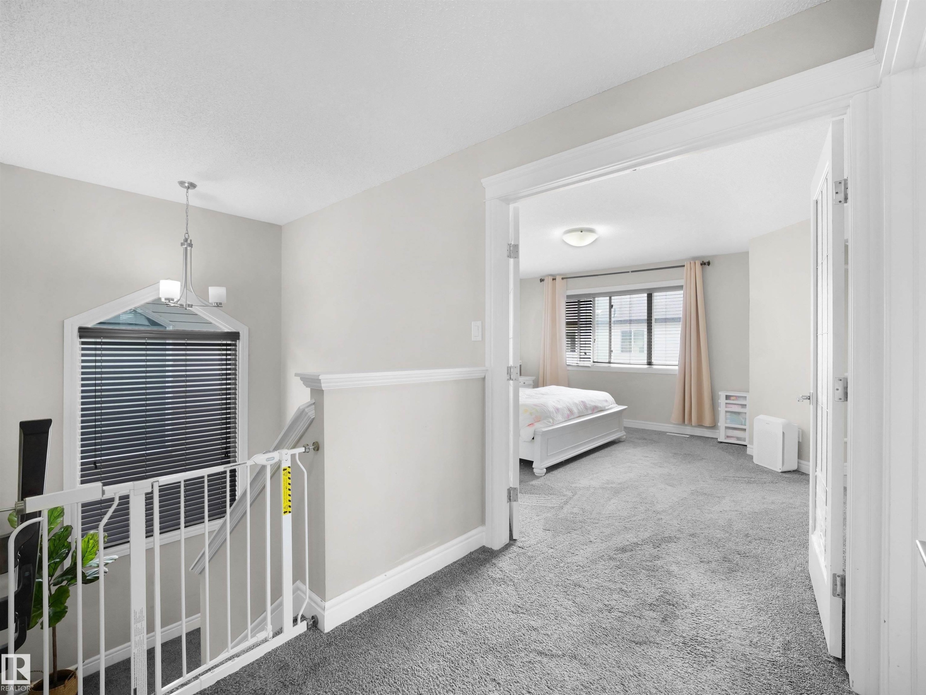 Carpeted hallway with white baseboards and trim, featuring a contemporary chandelier and an architectural window with blinds - 3247 15 Avenue Nw, Edmonton, AB - Indoor