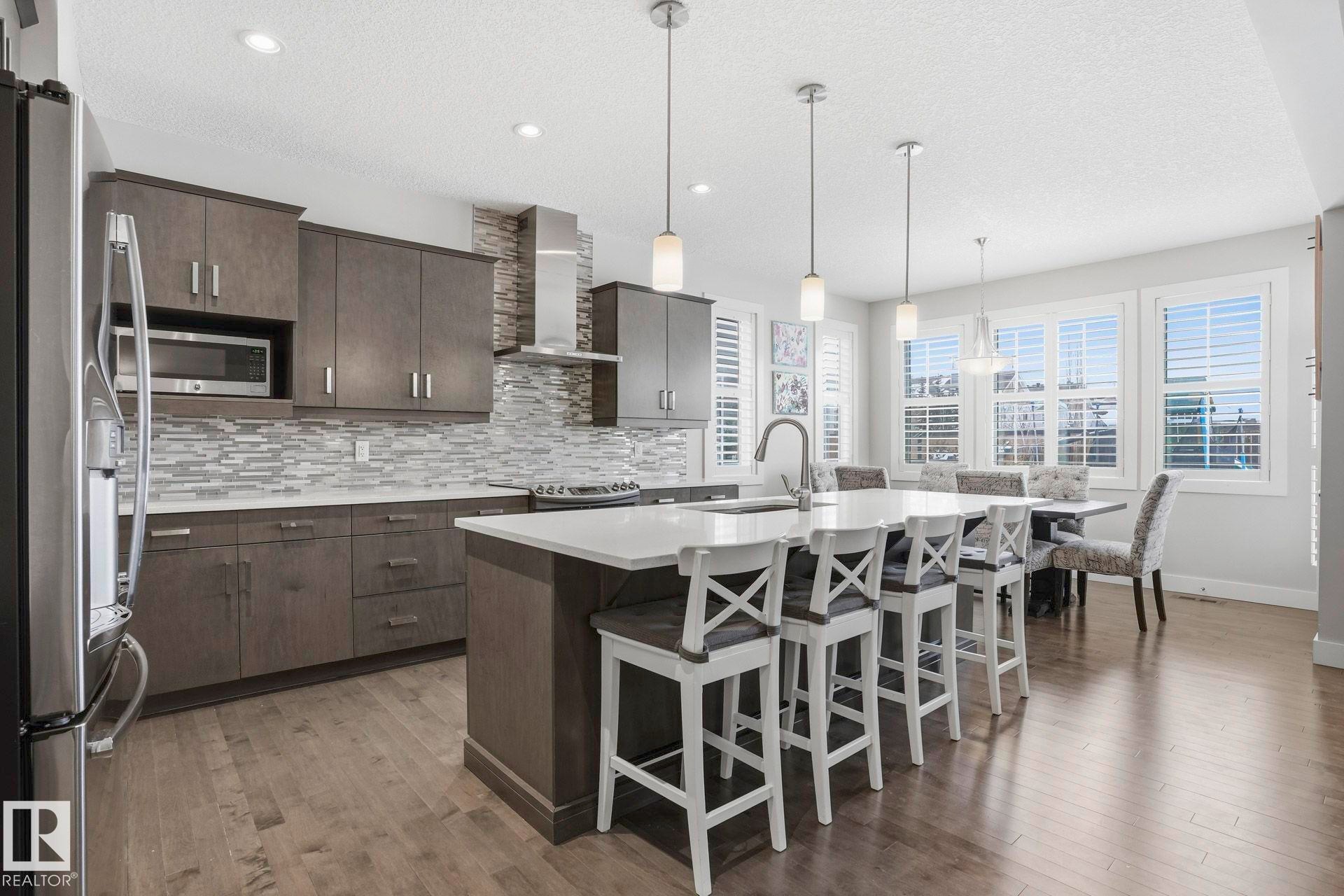 Modern kitchen featuring dark wood-finish cabinetry, stainless steel appliances, a tile backsplash, a large center island with seating, and wood-finish flooring - 1915 77 Street, Edmonton, AB - Indoor Photo Showing Kitchen With Upgraded Kitchen