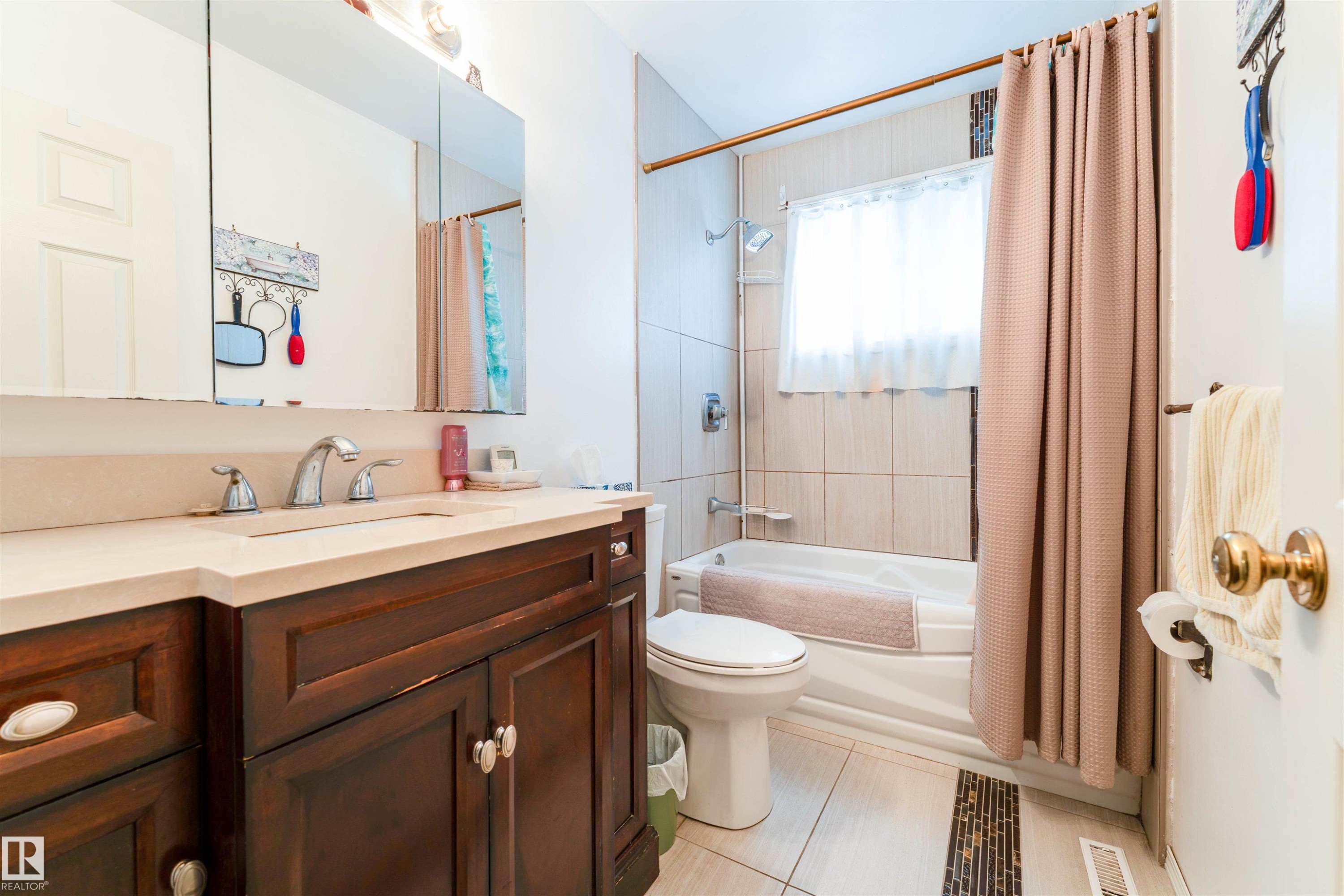 Bathroom featuring a rich wood-finish vanity with an integrated sink and light-toned countertop, a mirrored medicine cabinet, a tiled shower-tub combination, and tiled flooring with an accent strip - 13231 74 Street, Edmonton, AB - Indoor Photo Showing Bathroom