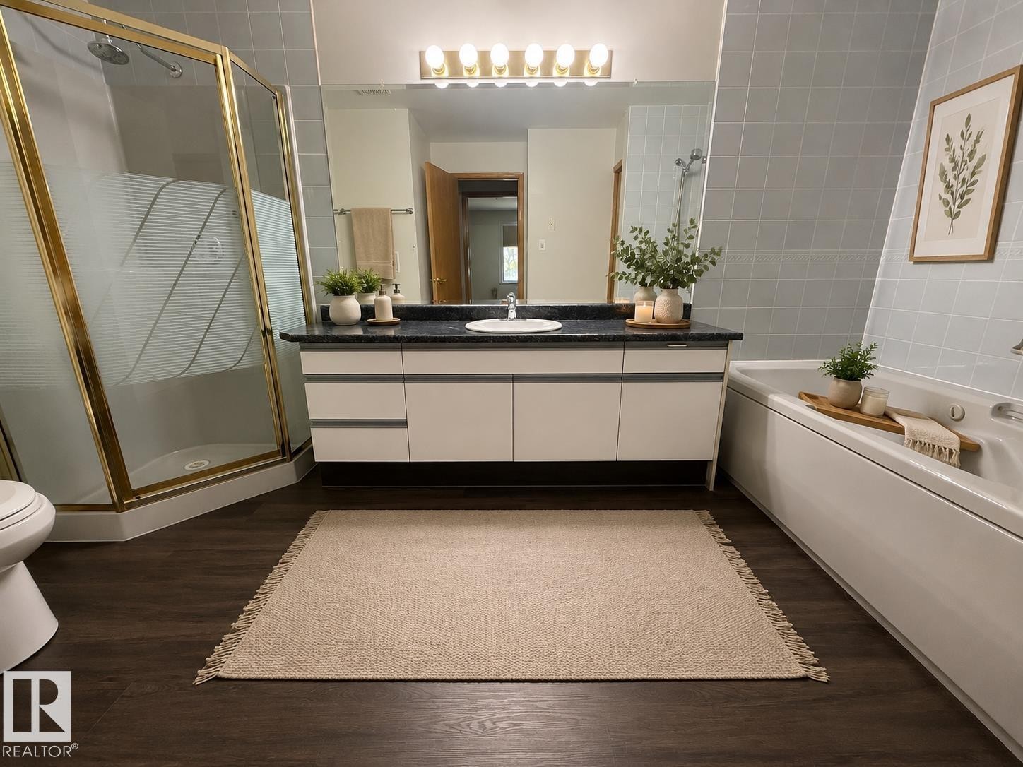 Bathroom featuring a dark wood-finish floor, a gold-framed glass shower enclosure, a vanity with white cabinetry and dark countertop, a mirror with sconce lighting, and a white soaking tub with tiled surround - #309 10610 76 St Nw, Edmonton, AB - Indoor Photo Showing Bathroom