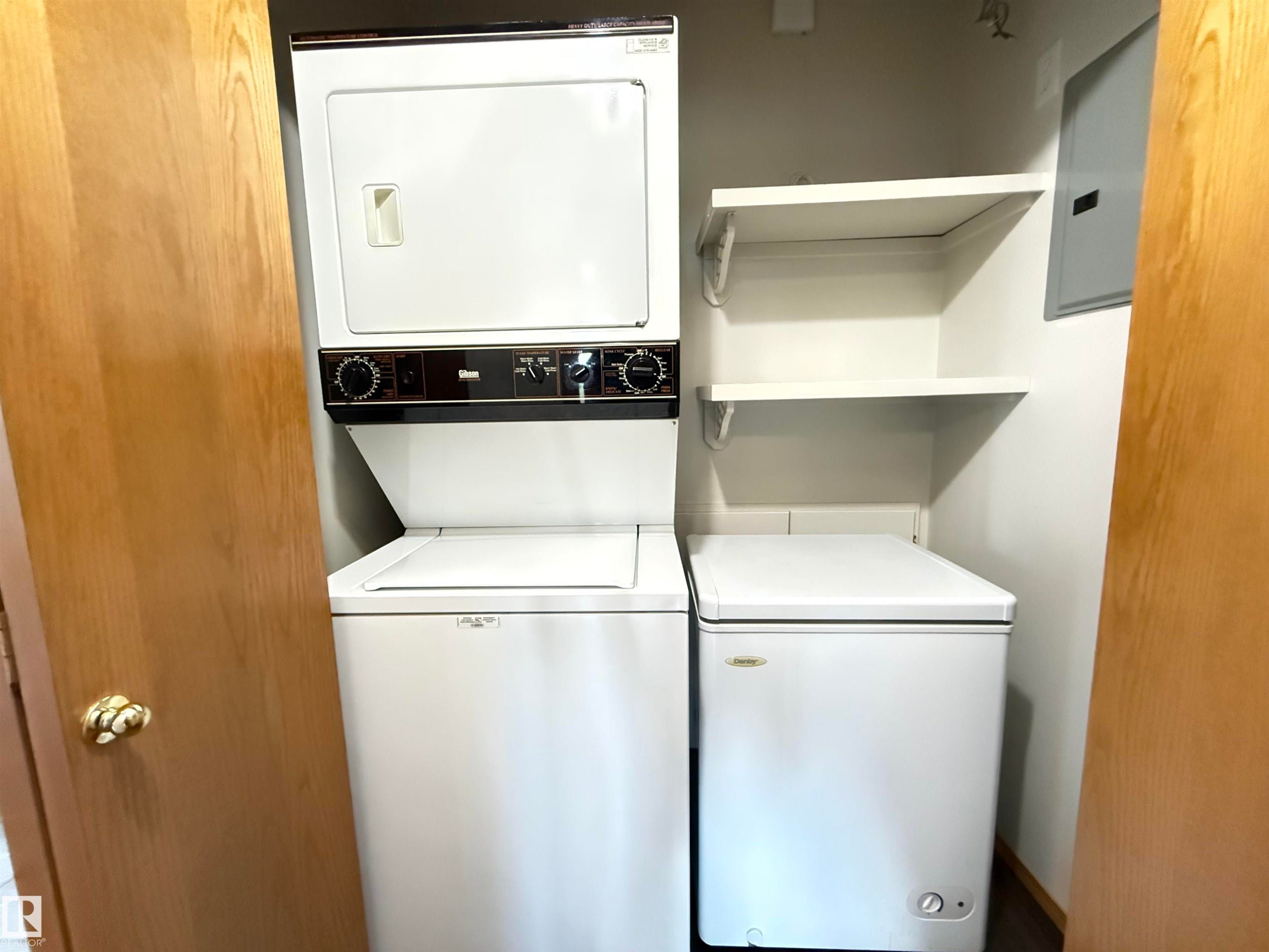 Dedicated utility closet featuring a stacked washer and dryer, additional chest freezer, and two white shelving units - #309 10610 76 St Nw, Edmonton, AB - Indoor Photo Showing Laundry Room