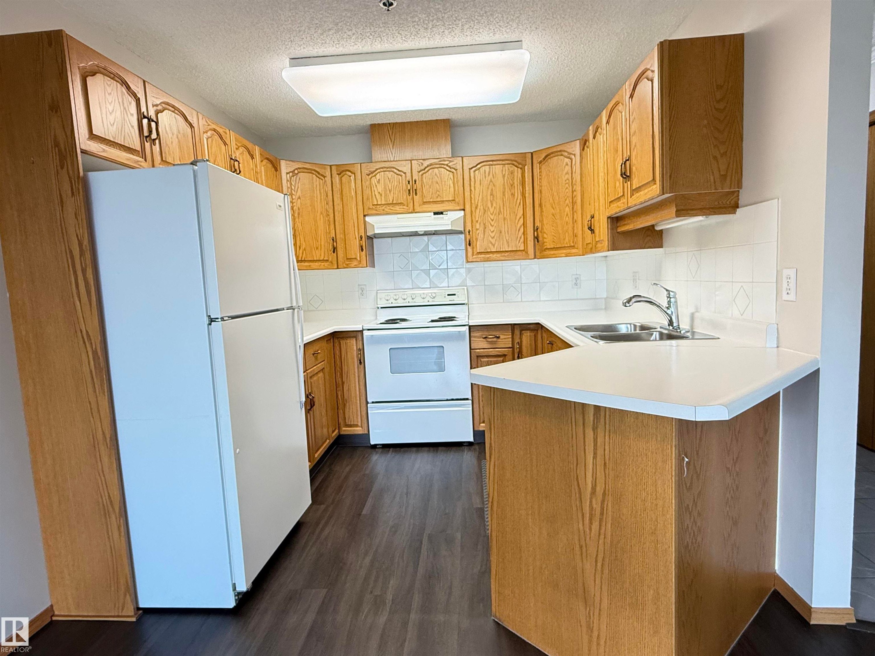 Kitchen featuring wood-finish cabinetry, white countertops, and white tile backsplash - #309 10610 76 St Nw, Edmonton, AB - Indoor Photo Showing Kitchen With Double Sink