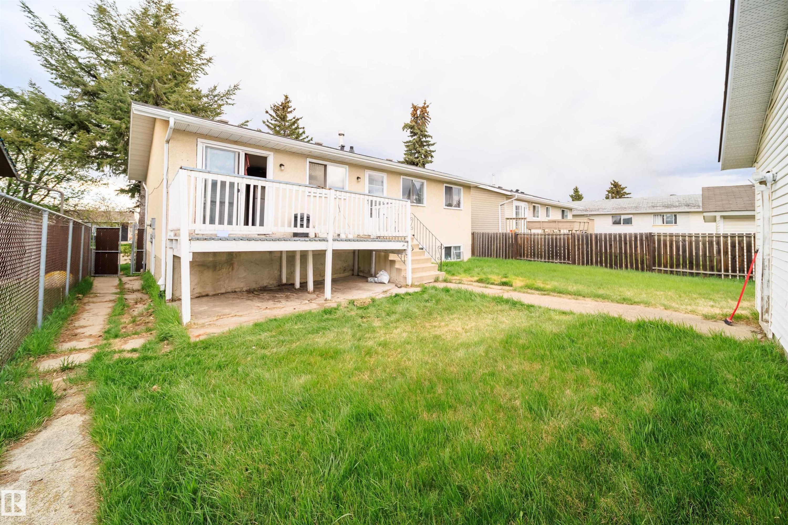 Expansive backyard featuring a raised wooden deck with white railing, a concrete patio area below the deck, a chain-link fence bordering a side pathway, and a wooden privacy fence at the property's rear - 1771 37 Street, Edmonton, AB - Outdoor With Deck Patio Veranda