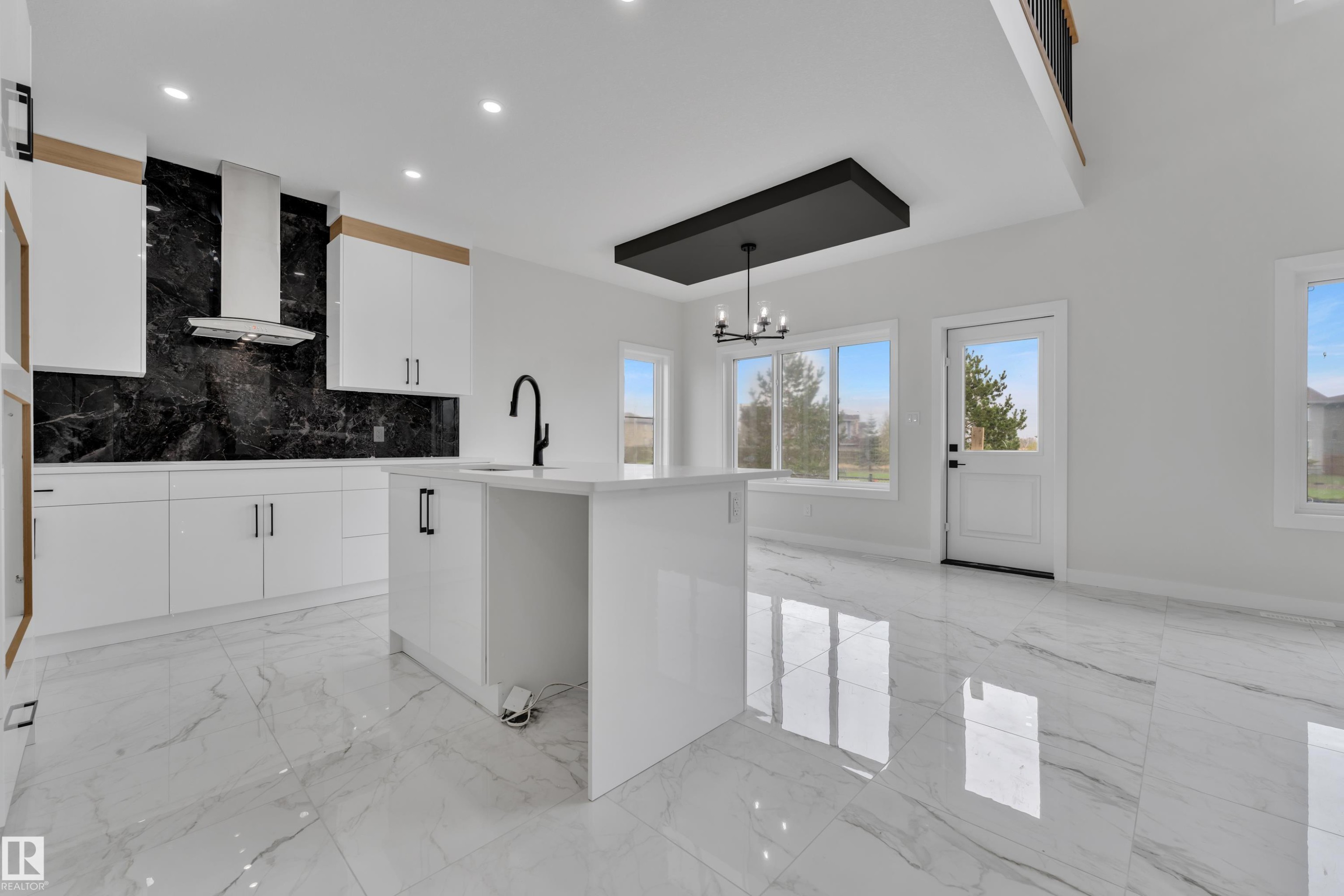 Modern kitchen and living area featuring a large central island, white cabinetry with wood accents, a black slab backsplash, stainless steel range hood, and polished tile flooring throughout - 12623 44 Street, Edmonton, AB