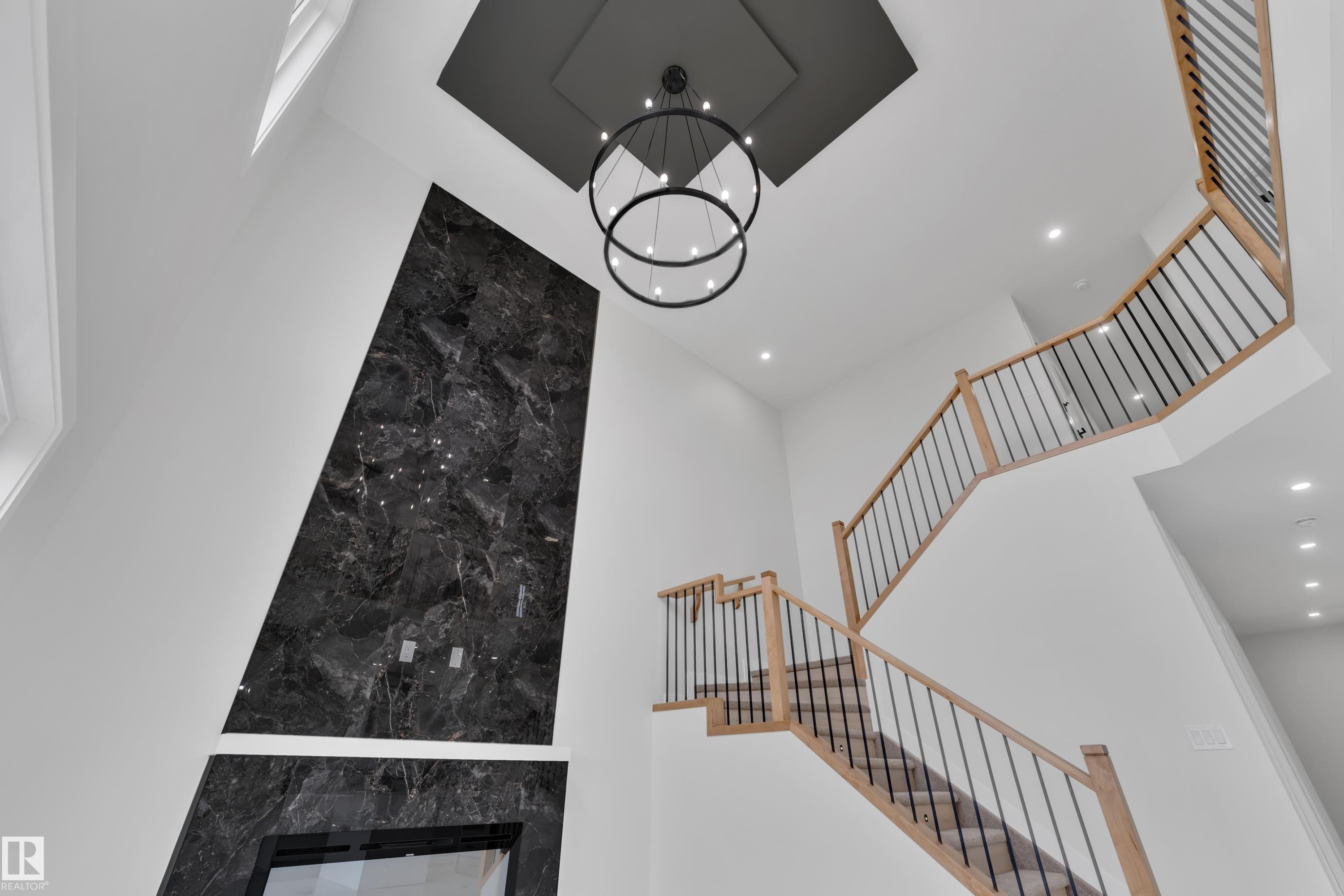 Two-story foyer featuring a striking dark marble-effect fireplace surround, a prominent contemporary chandelier, and a custom wood-finish staircase with black metal balusters - 12623 44 Street, Edmonton, AB