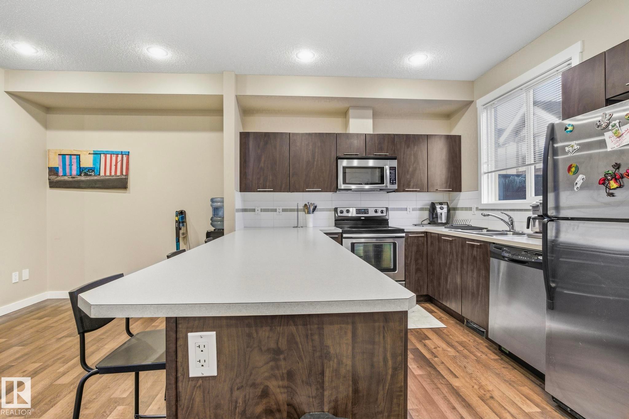 Contemporary kitchen featuring wood-finish cabinetry, stainless steel appliances, a large island with white countertops, and wood-finish flooring - 30 6075 Schonsee Way, Edmonton, AB - Indoor Photo Showing Kitchen With Stainless Steel Kitchen