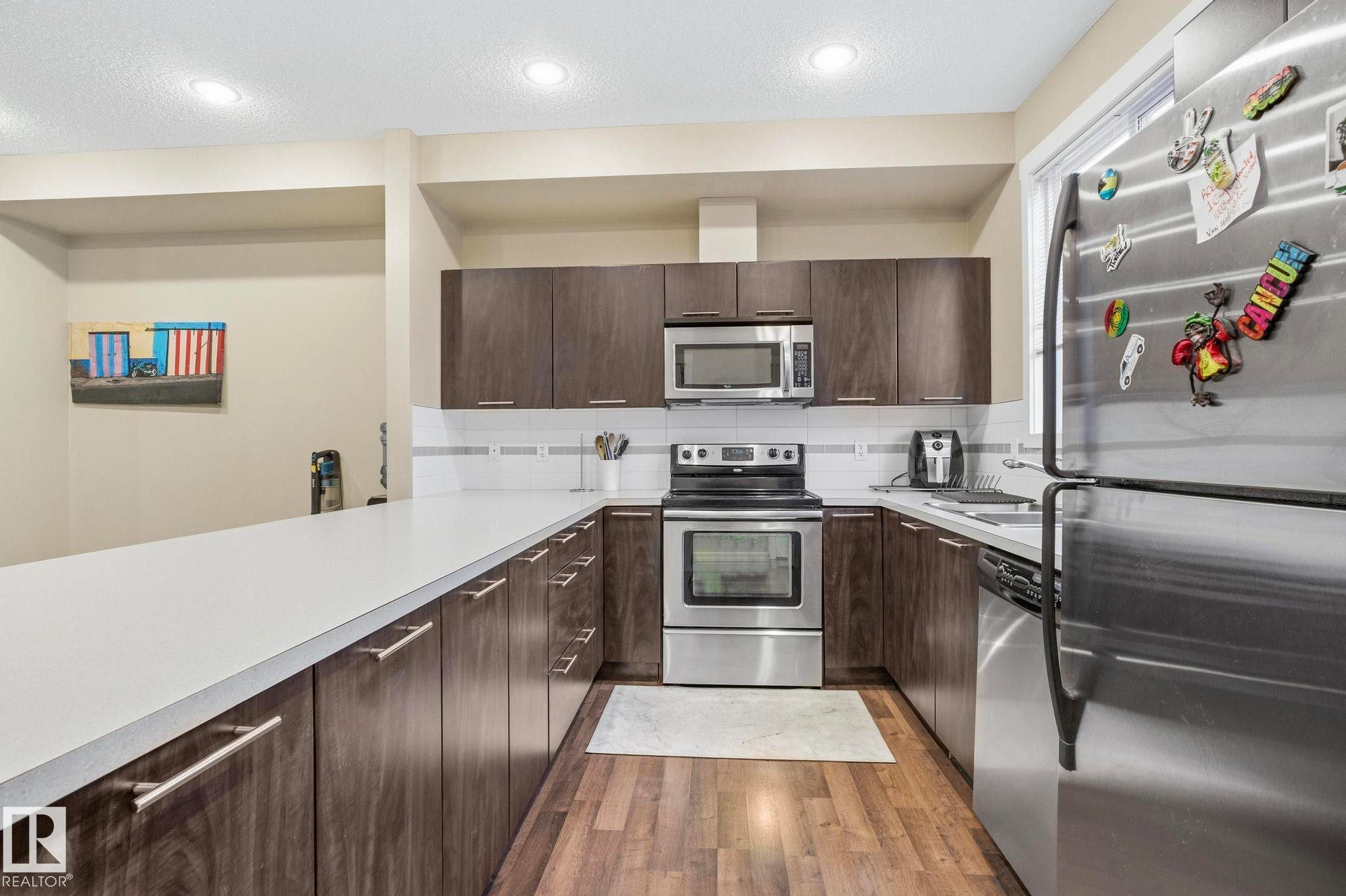 Modern kitchen featuring dark wood-finish cabinetry, light-toned countertops, a full suite of stainless steel appliances, and wood-finish flooring - 30 6075 Schonsee Way, Edmonton, AB - Indoor Photo Showing Kitchen With Stainless Steel Kitchen With Upgraded Kitchen