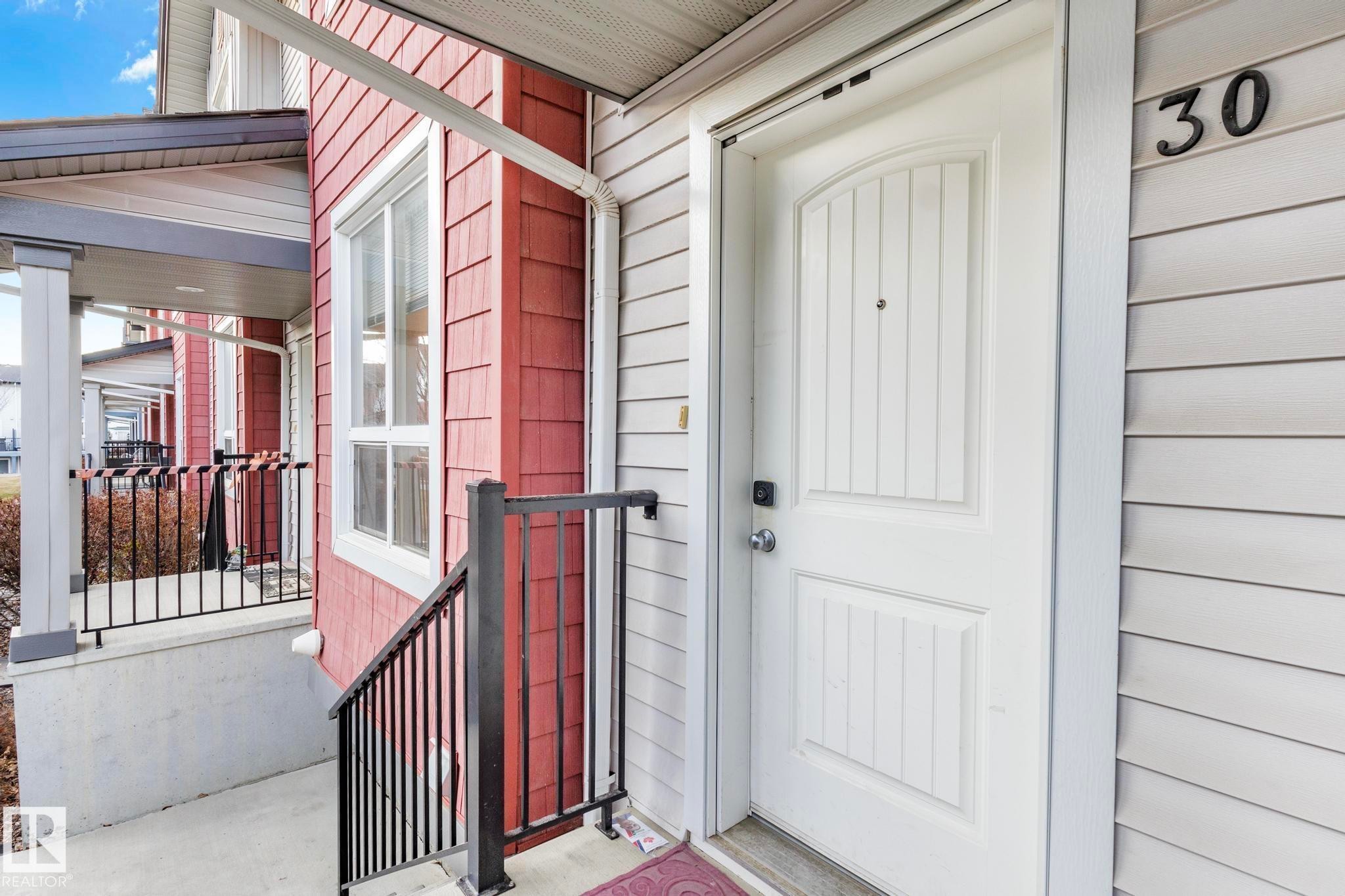 Entryway featuring a white paneled door with integrated house number, light-colored siding, and a red shingle accent wall - 30 6075 Schonsee Way, Edmonton, AB - Outdoor With Exterior